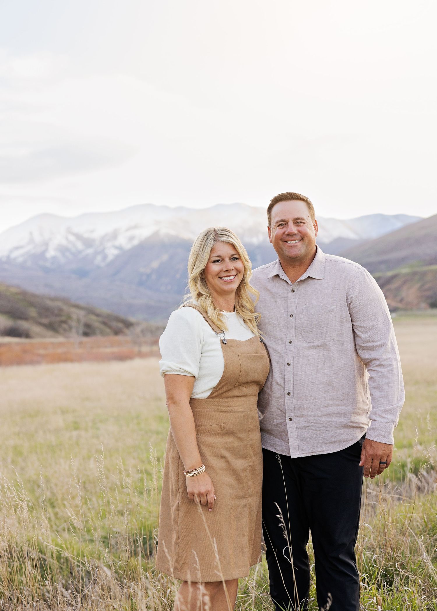 A smiling couple stands in a mountain meadow with snow-capped peaks behind them, woman in tan dress, man in gray shirt.