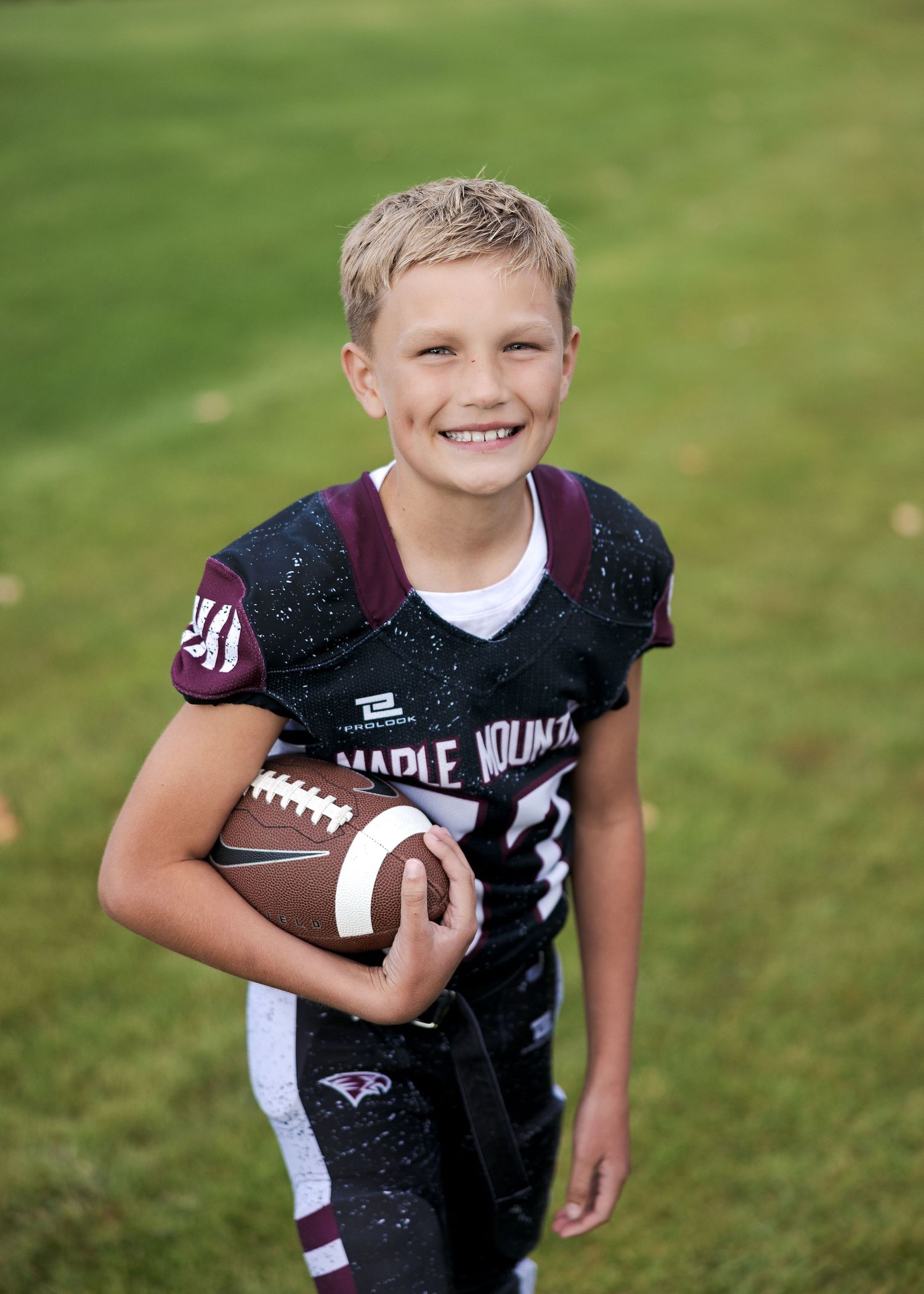 Young football player in black uniform holds ball while posing on grass field during sports photo session.