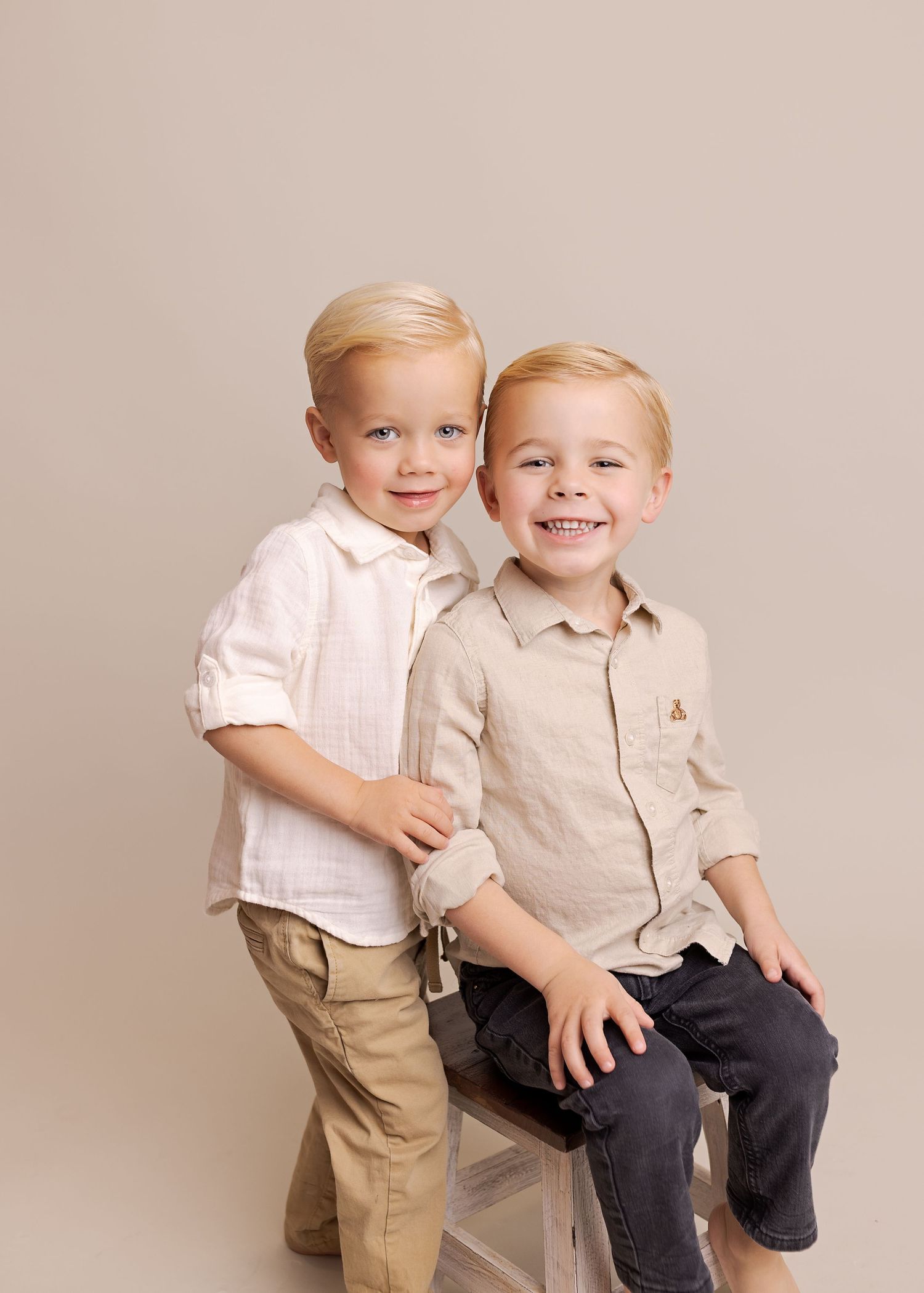 Two young children in light colored clothing playfully interact during a photo session against a neutral background.