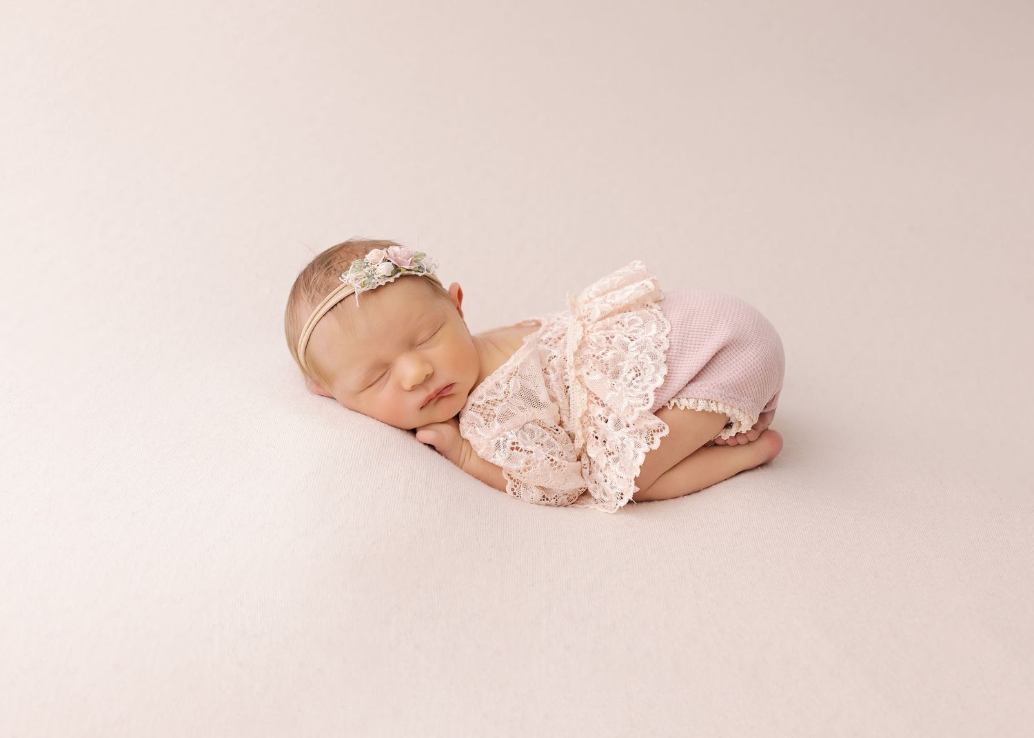 Sleeping newborn in delicate pink lace outfit during professional photo session.