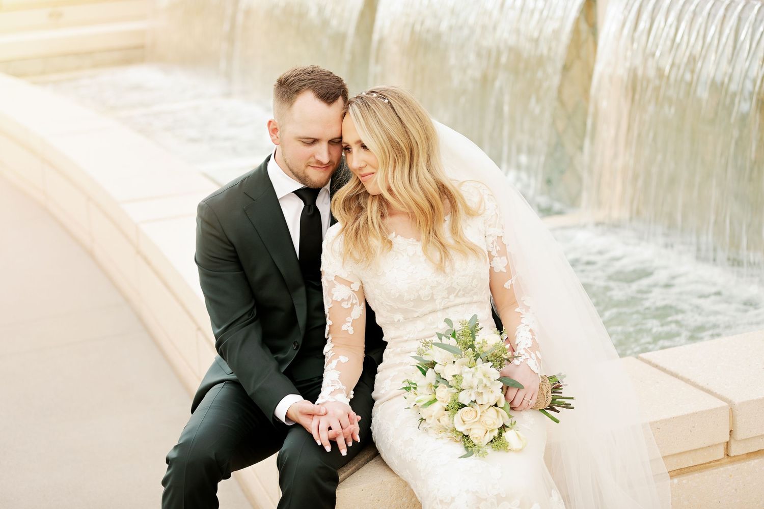 Couple in wedding attire sitting by water feature.
