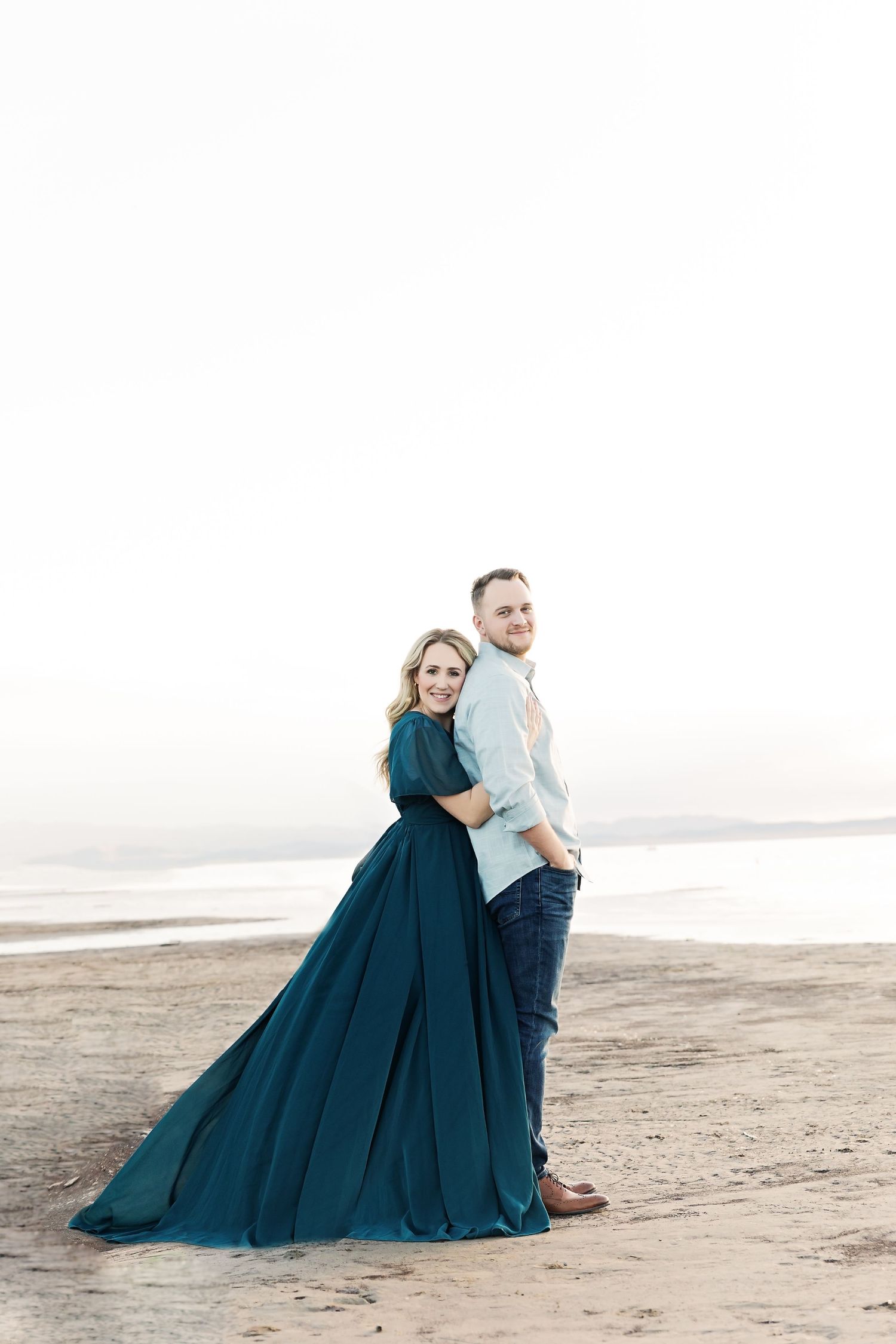 A couple in formal wear poses back-to-back on a beach, with the woman wearing a flowing teal gown.