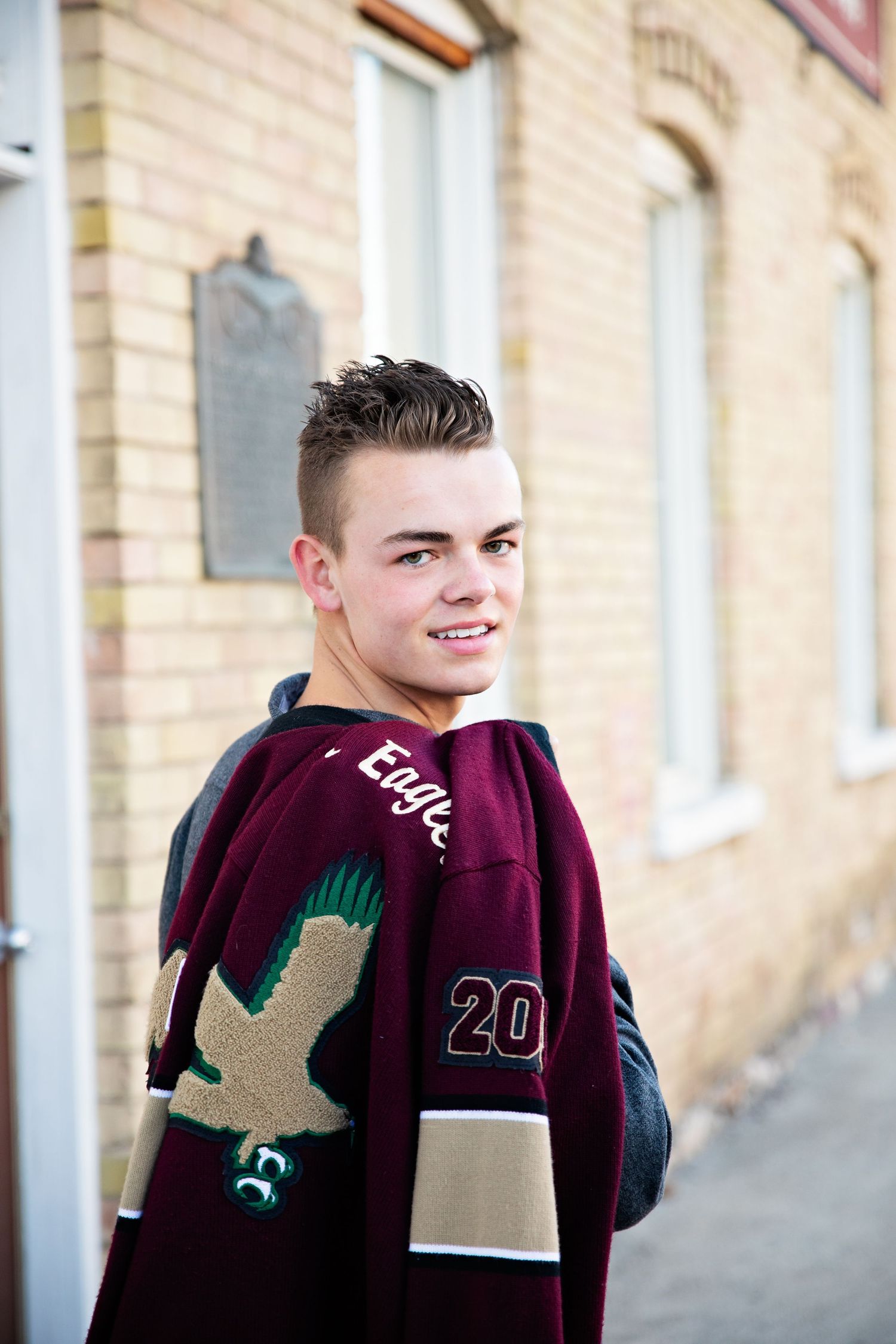 Athletic portrait wearing maroon varsity jacket with eagle emblem against brick building.