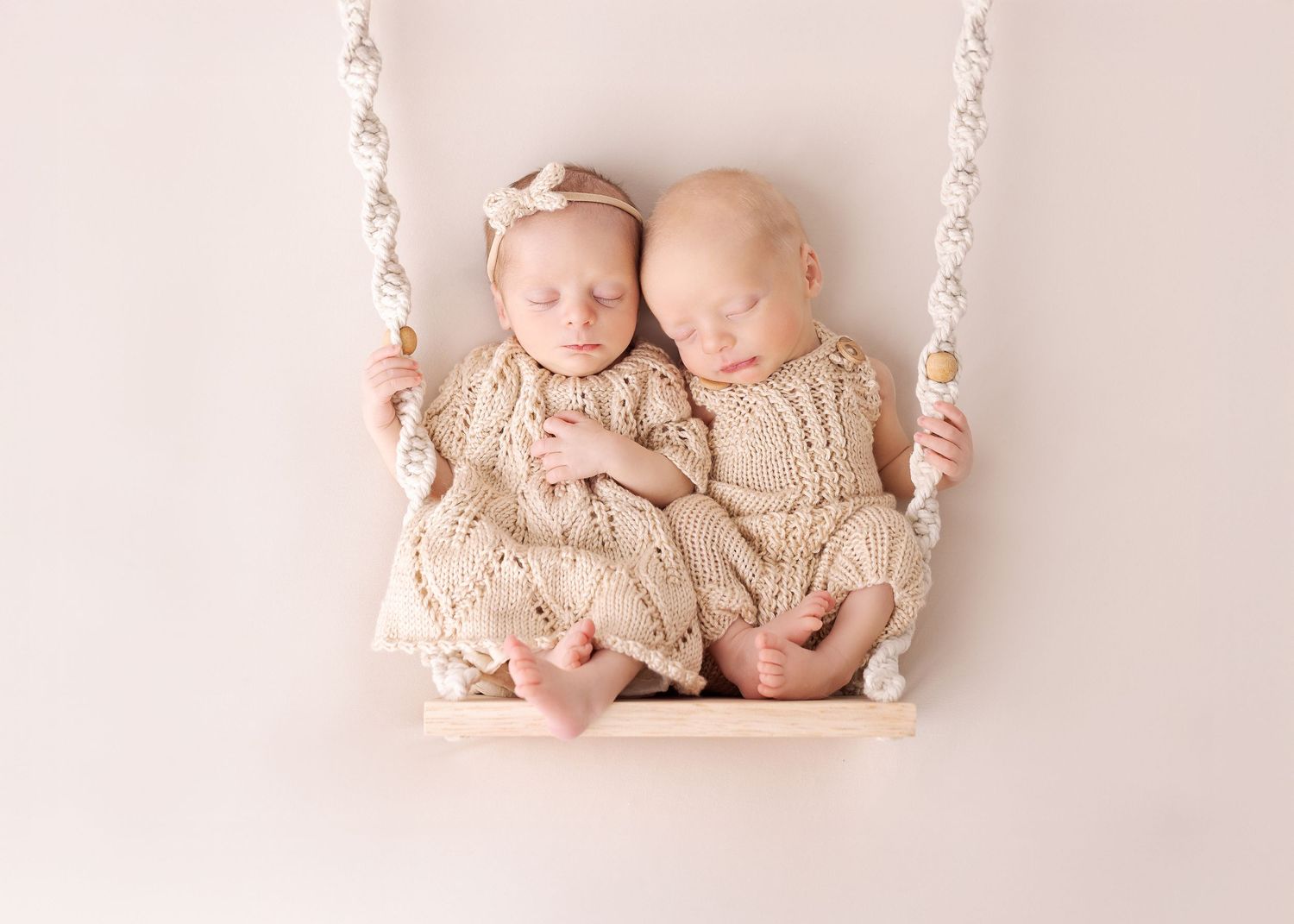 Two sleeping babies in matching cream knit outfits sit together on a small wooden swing with white beaded ropes.