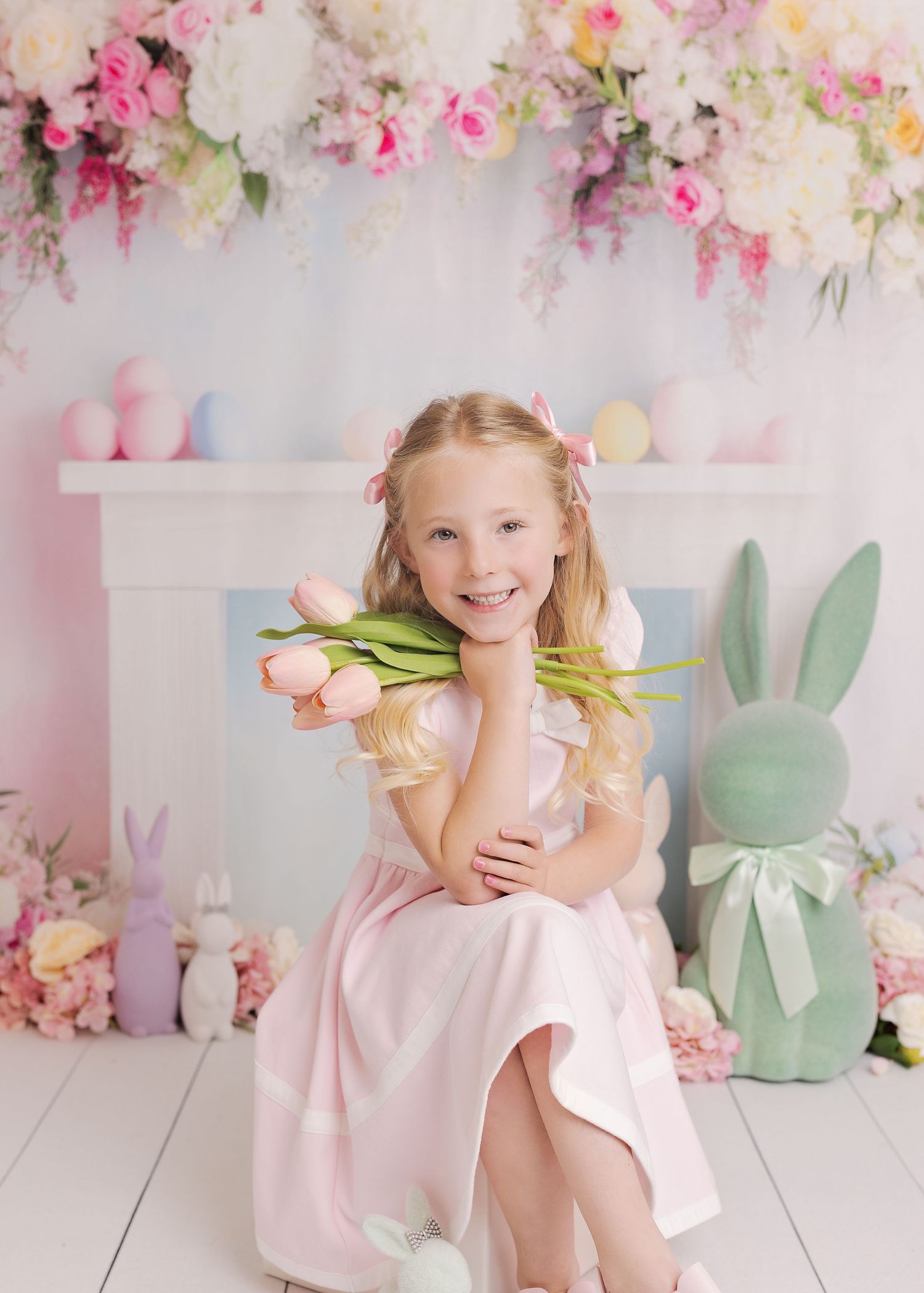 A child in a pink dress poses with tulips against a floral backdrop with mint green bunny decorations.