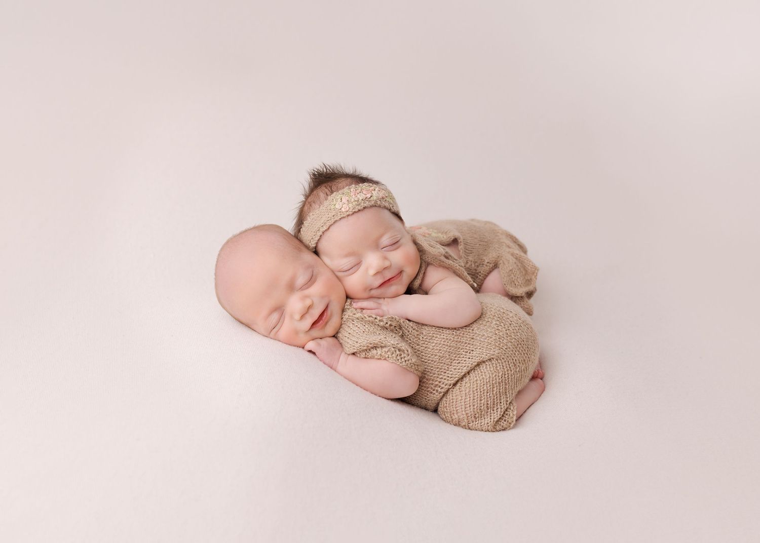 Sleeping newborn baby dressed in soft beige knit outfit lying peacefully on cream backdrop during professional photo session.