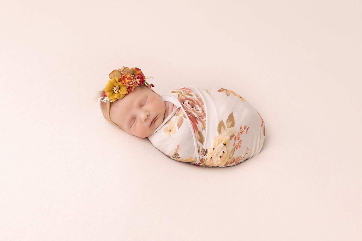 Sleeping newborn wrapped in floral swaddle wearing delicate flower crown on pink background.