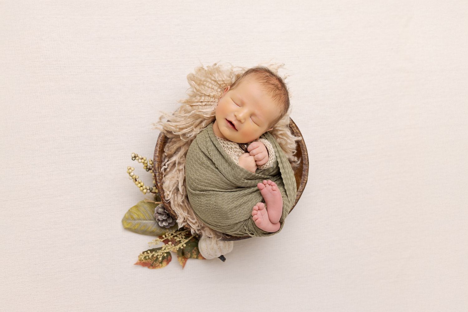 Sleeping baby wrapped in olive green swaddle nestled in rustic basket with dried flowers.
