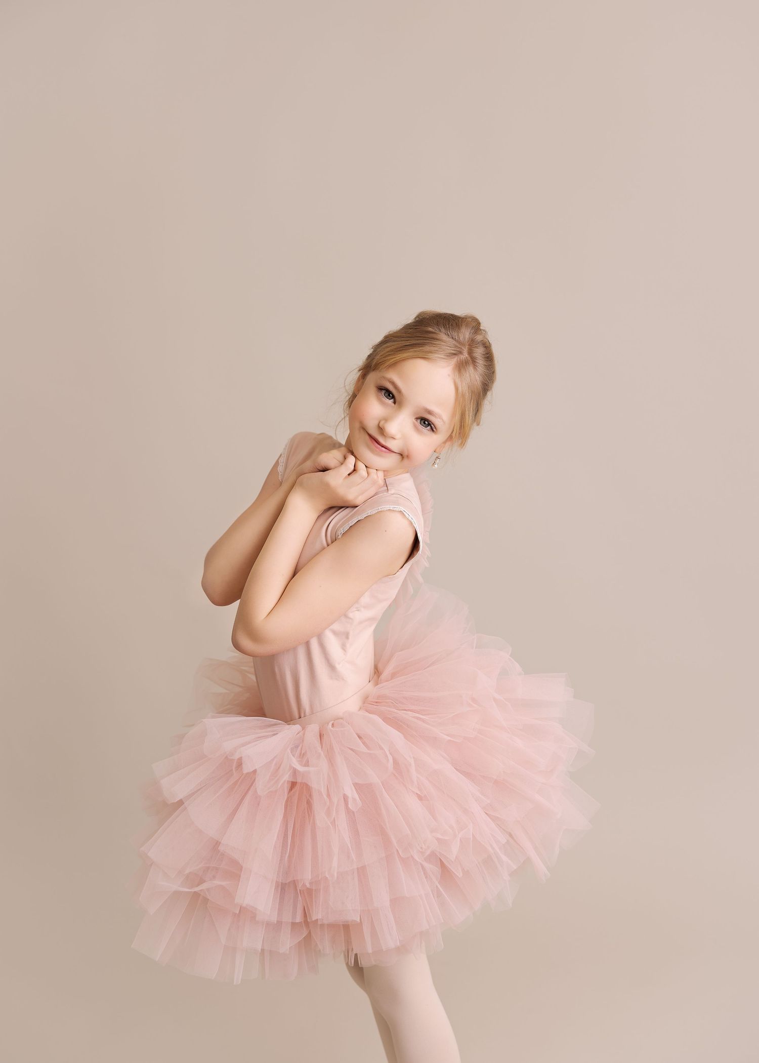 Ballet dancer in pink tulle tutu posing playfully against beige wall.