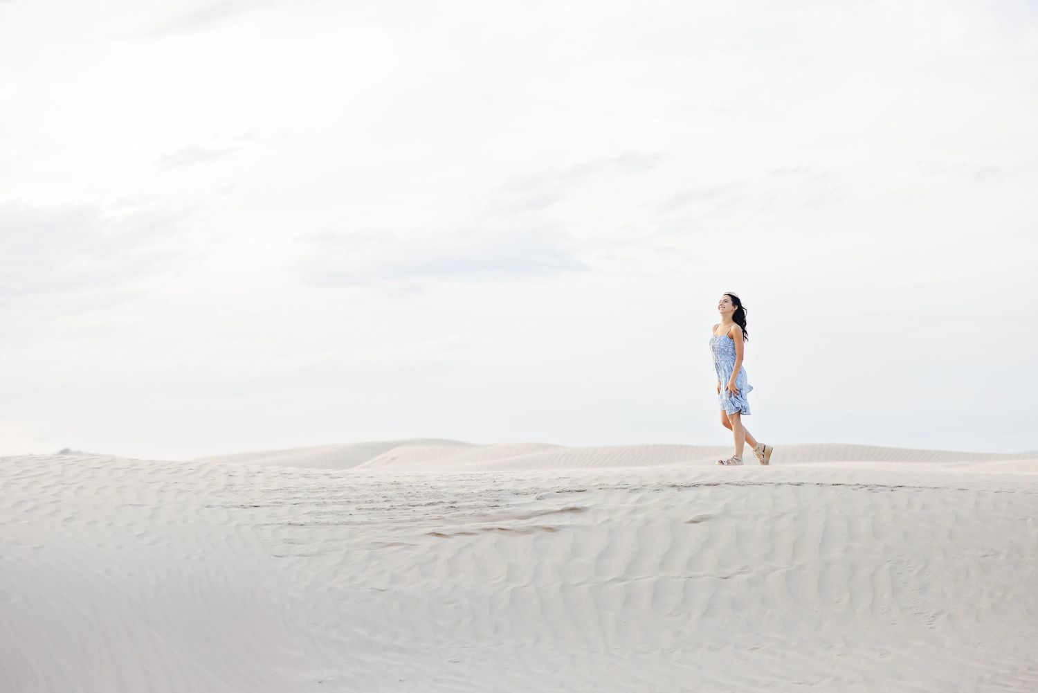 A lone figure in a blue dress walks along a pristine white sand dune against a bright overcast sky.