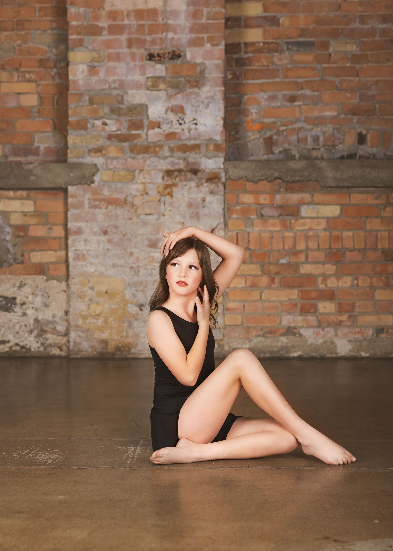 A dancer in a black leotard poses gracefully against a rustic brick wall backdrop in an artistic composition.