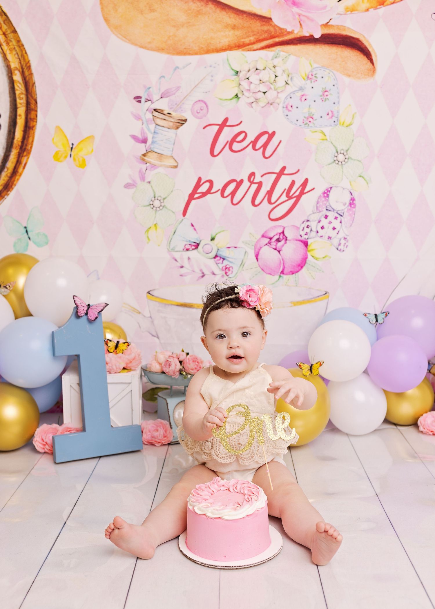 A baby in a sparkly gold dress sits near a pink cake and pastel decorations at a tea party themed first birthday celebration.