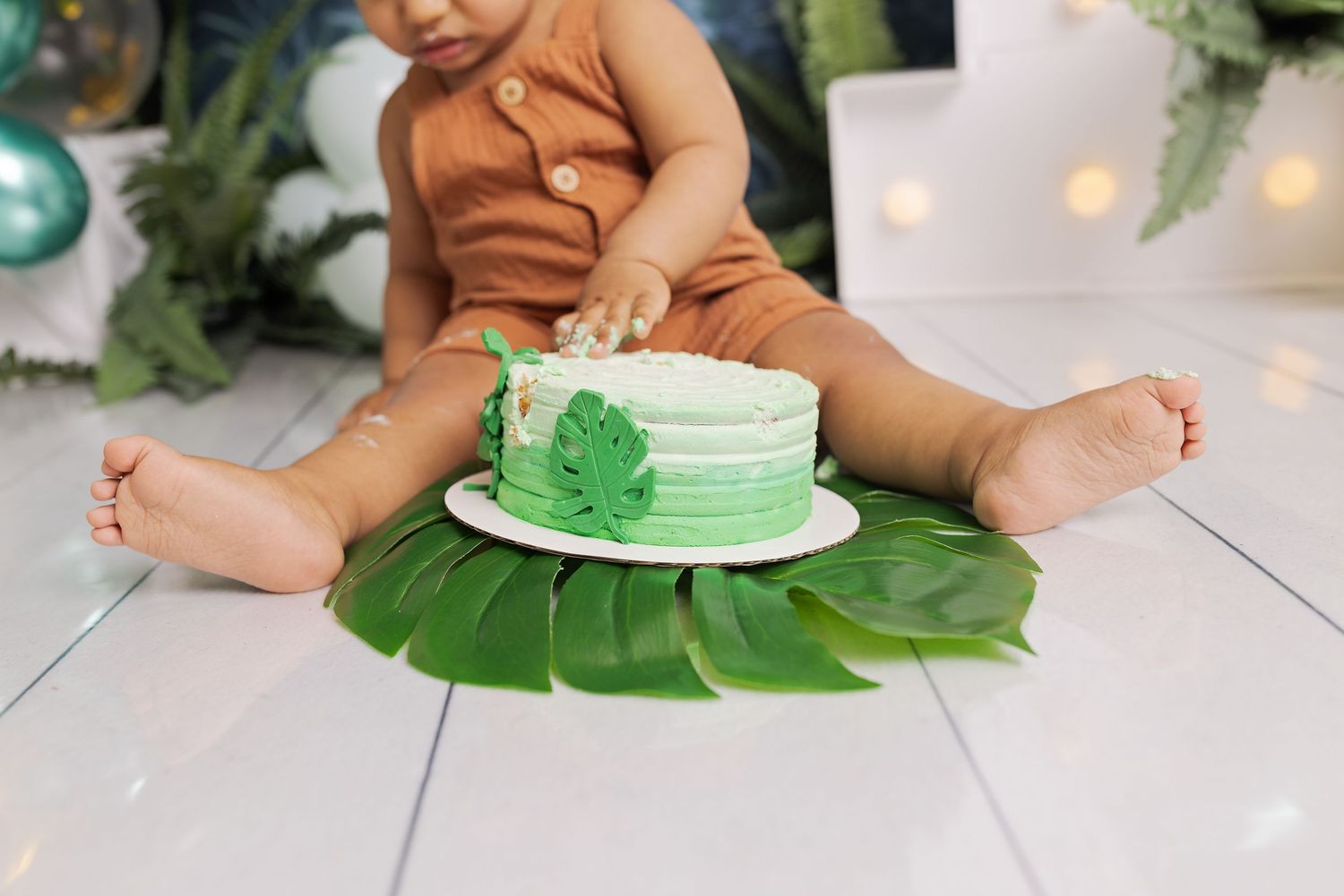 A baby in brown clothing smashes into a white and green frosted smash cake during a first birthday photoshoot.