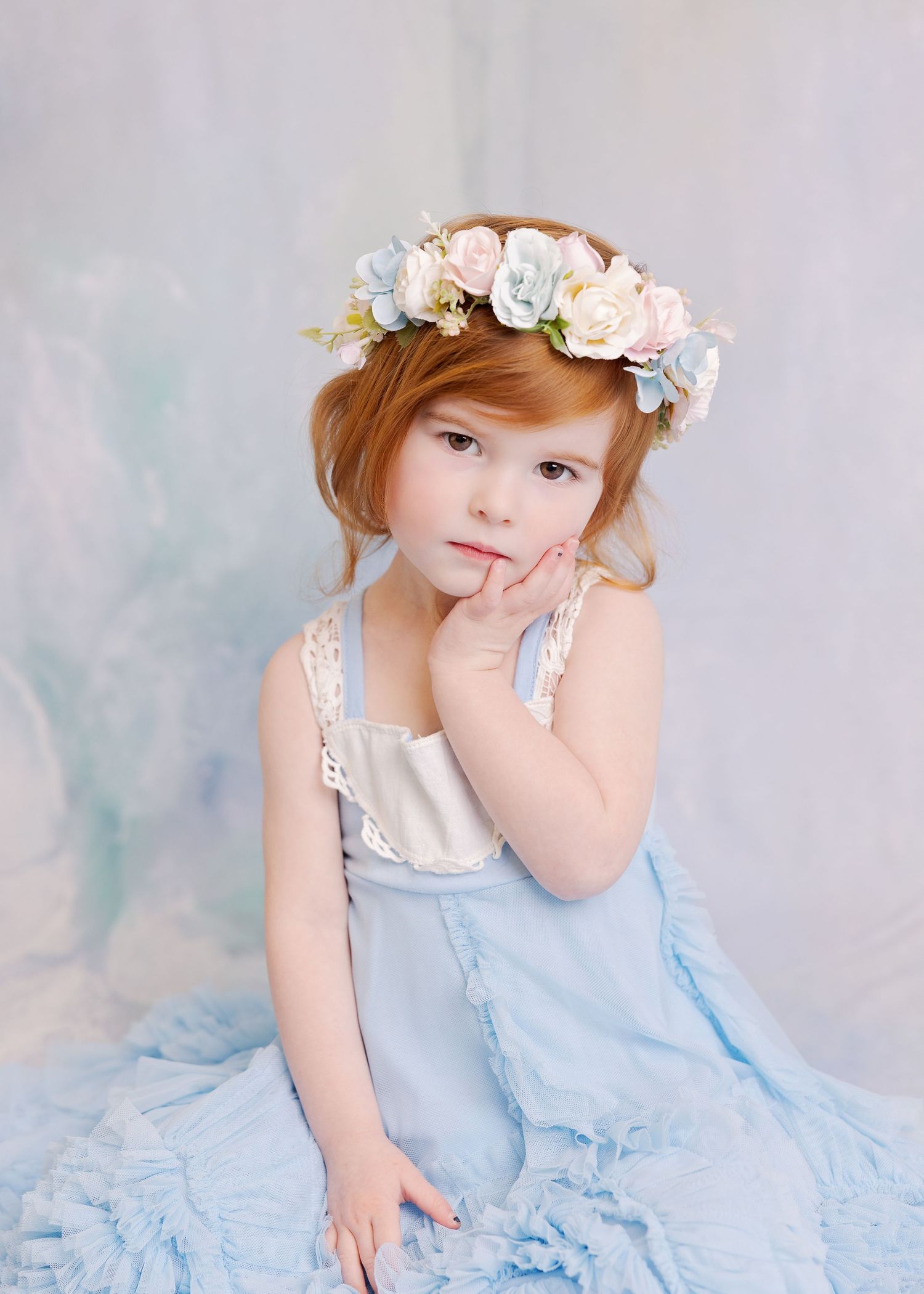 Ethereal portrait in light blue dress with floral crown against soft backdrop.