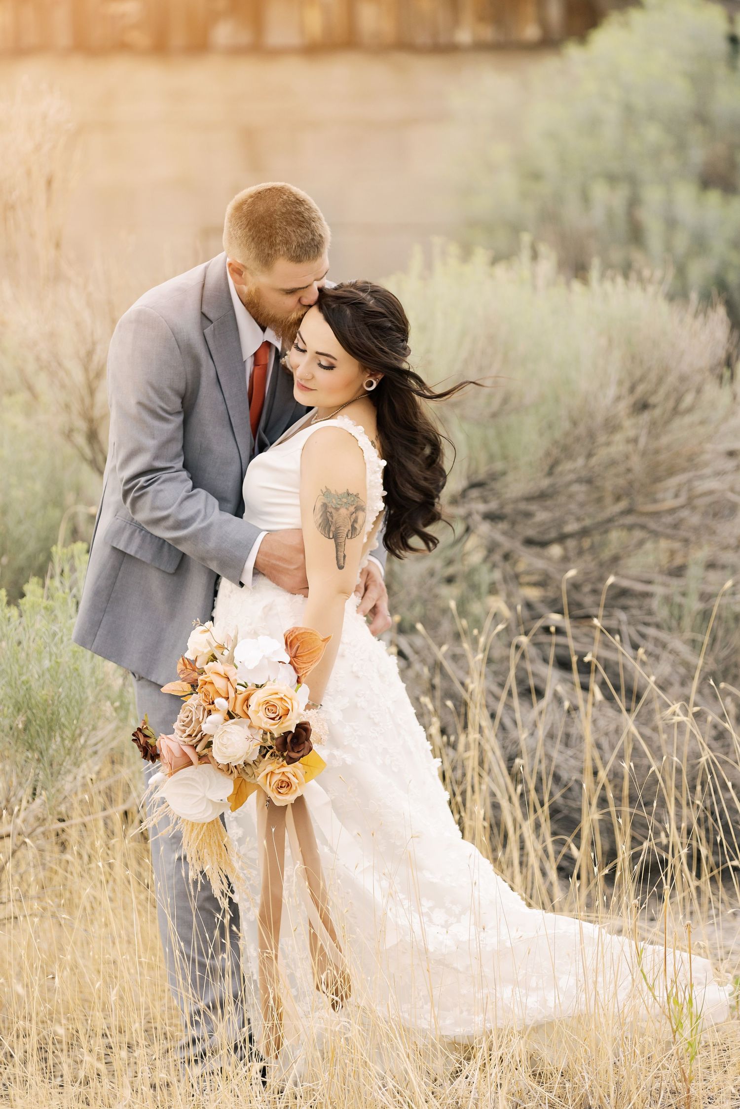 Romantic wedding couple embraces in field of sagebrush at sunset wearing grey suit and white dress.