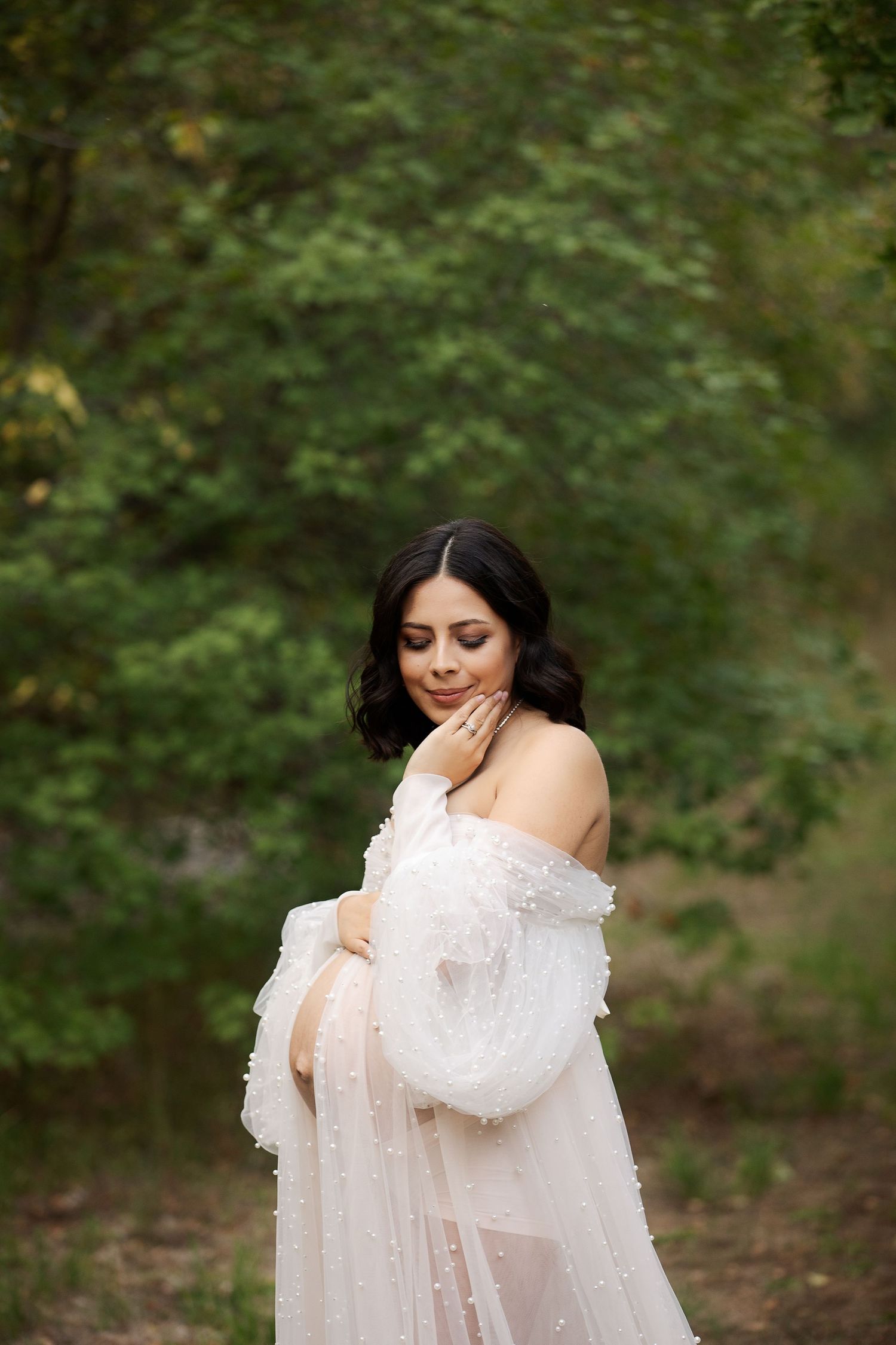 Maternity photo of woman in white lace dress standing in front of lush green forest.