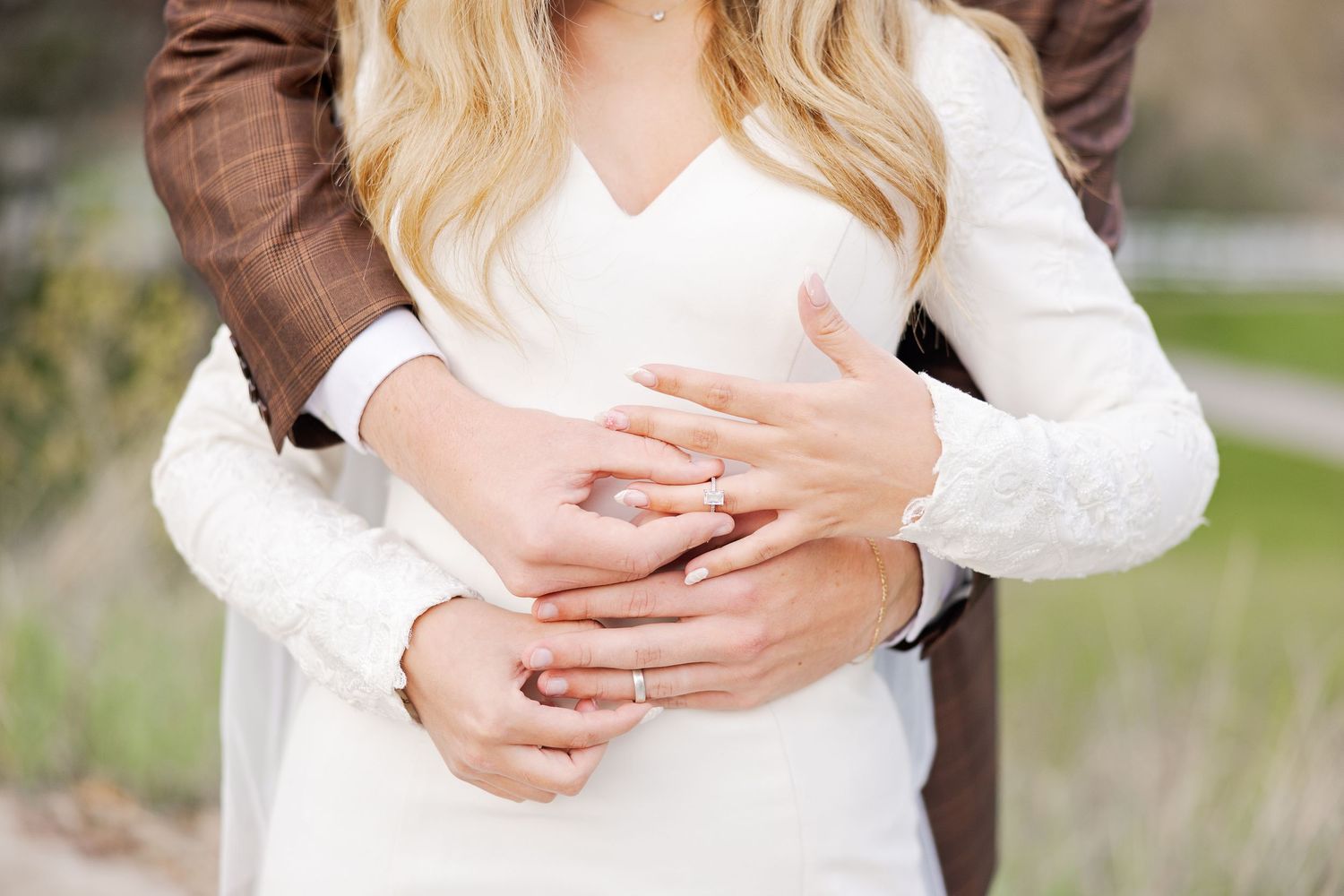 Close up of embracing couple showing hands with engagement rings against white sweater in outdoor setting.