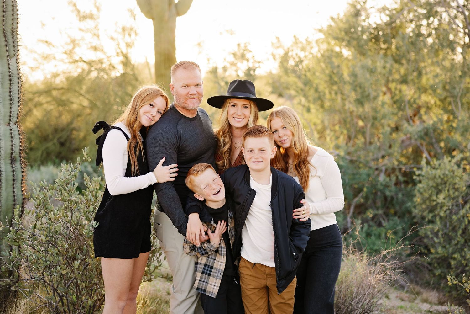 A casual family portrait taken against a desert landscape with saguaro cacti at sunset.