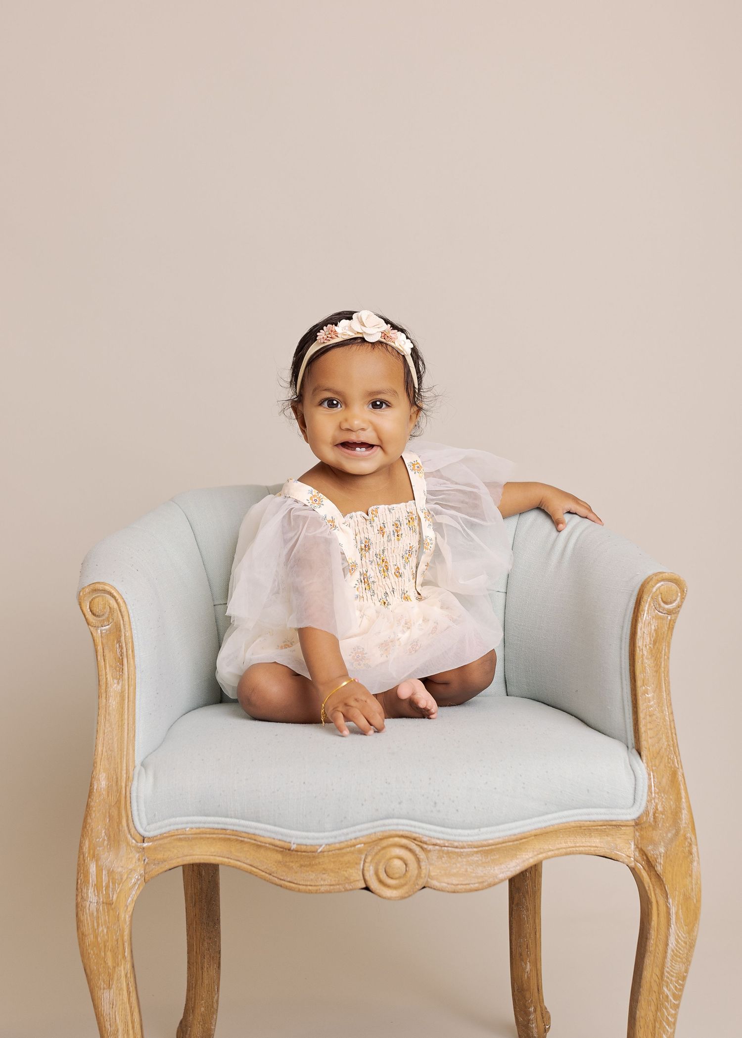 Smiling baby wearing white dress and floral headband seated on light blue antique chair.