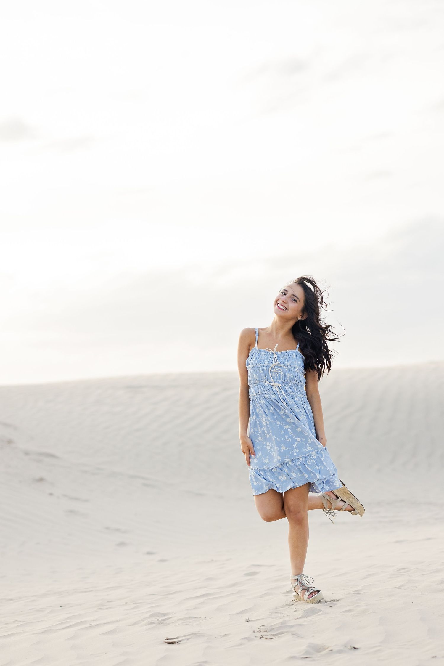 Woman in light blue summer dress walks joyfully across white sand dunes under cloudy sky.