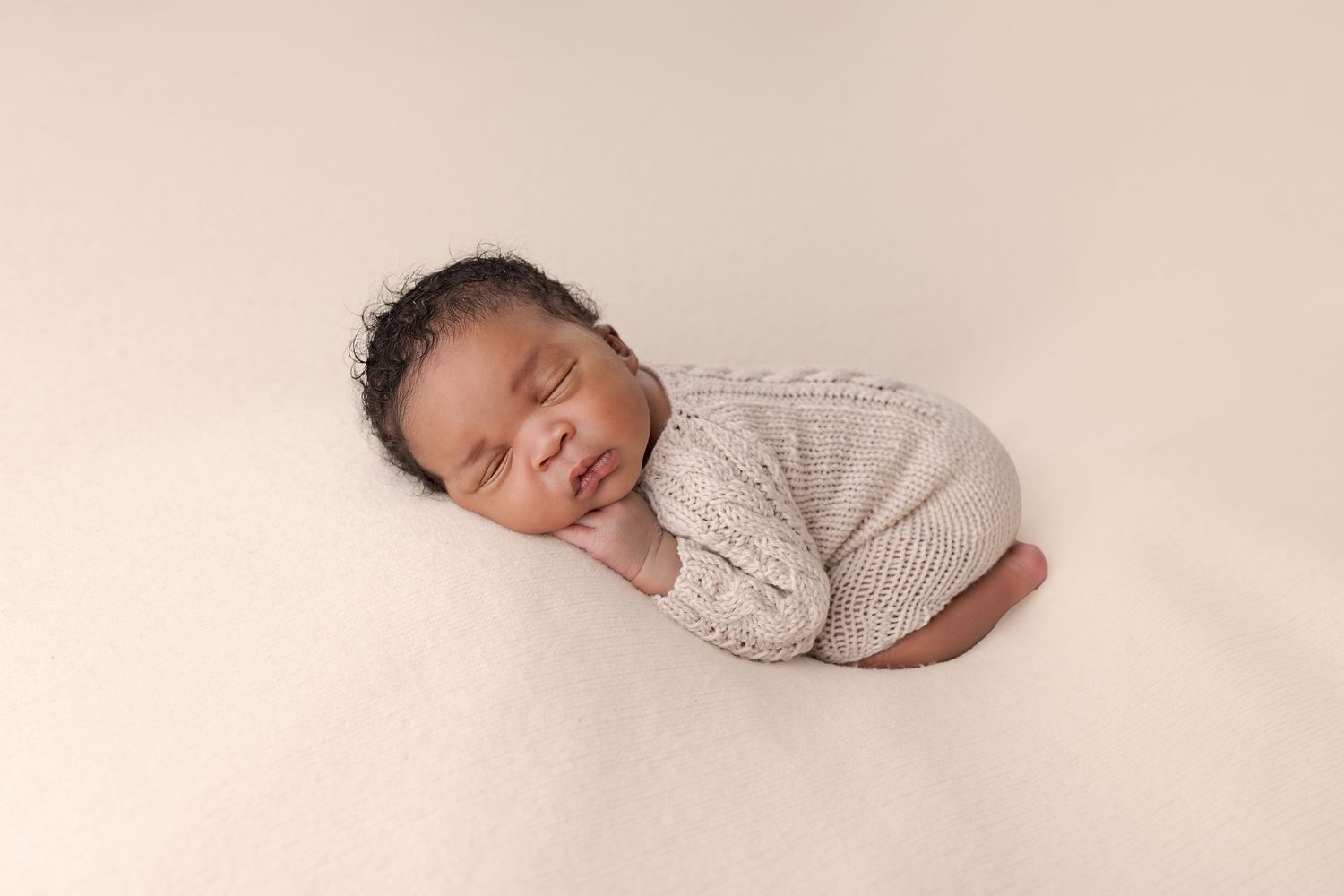 A sleeping newborn baby wrapped in soft white fabric lies peacefully against a neutral beige background.