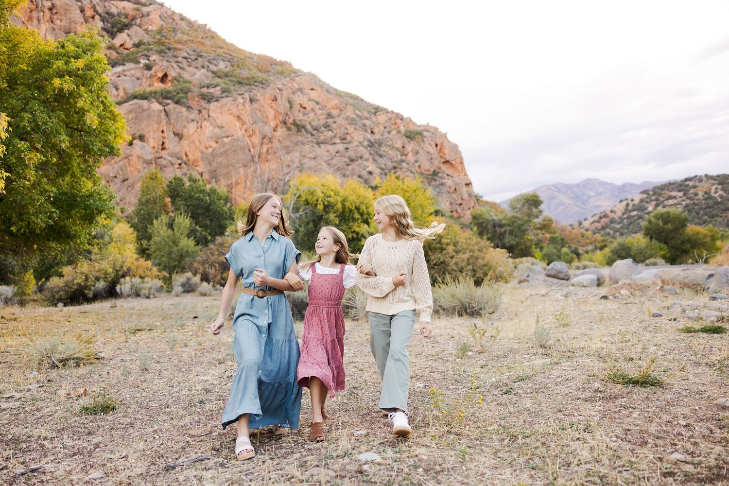 Three women walking together through a scenic desert landscape with rocky mountains and green trees in the background.