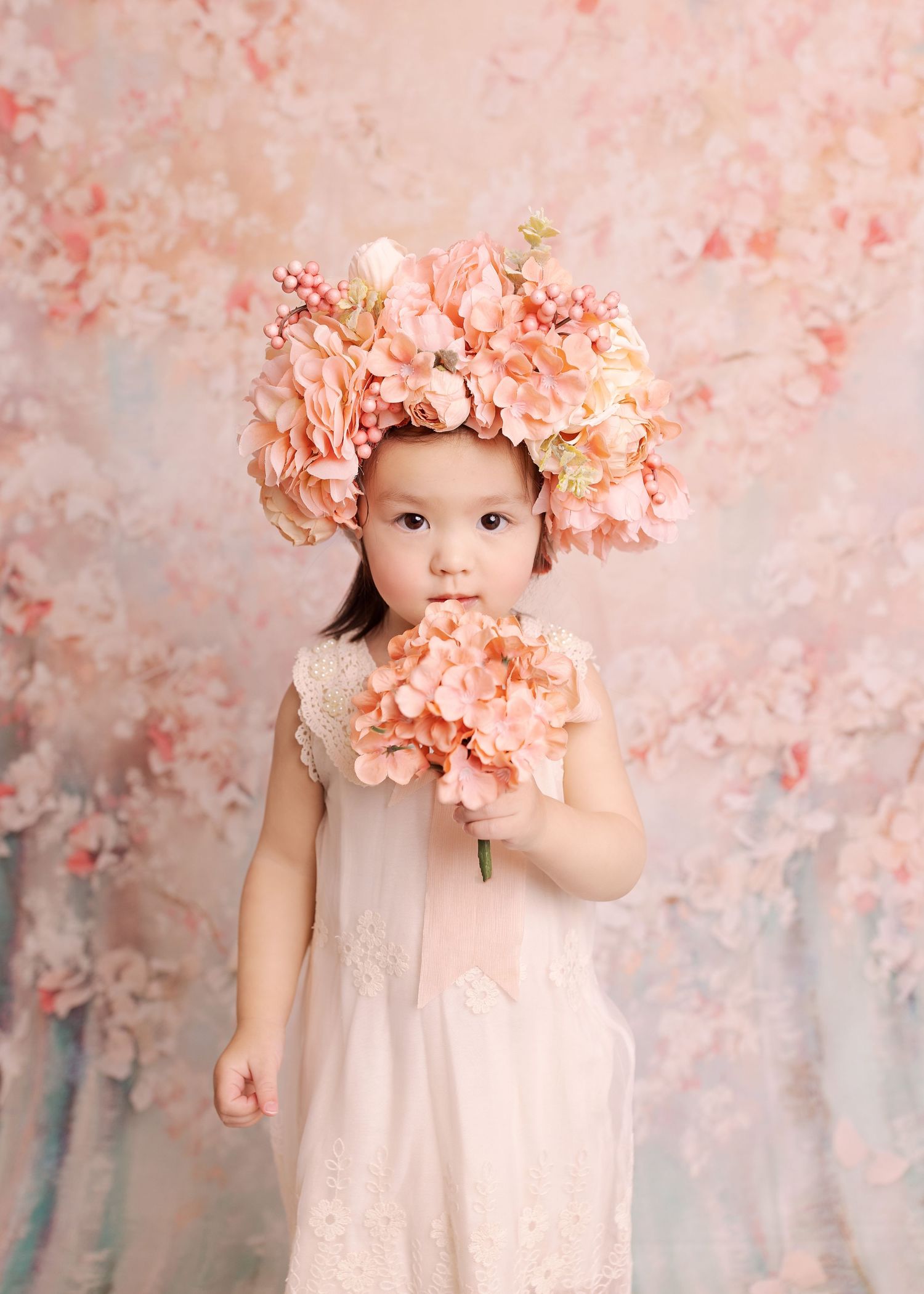 Young child in pink floral crown and dress posed against peachy pink floral backdrop.