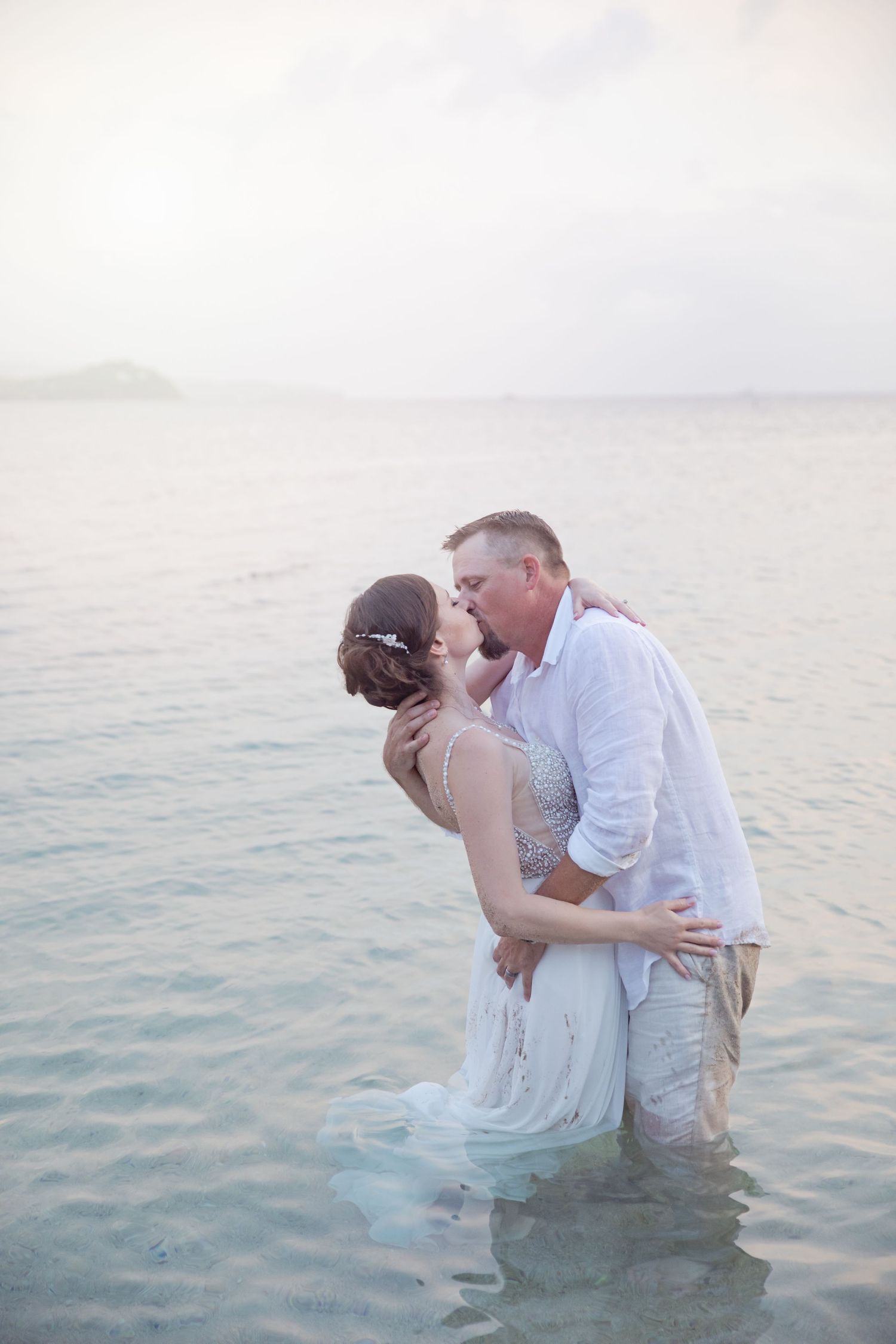 A romantic couple kissing while standing in shallow ocean water at dusk.