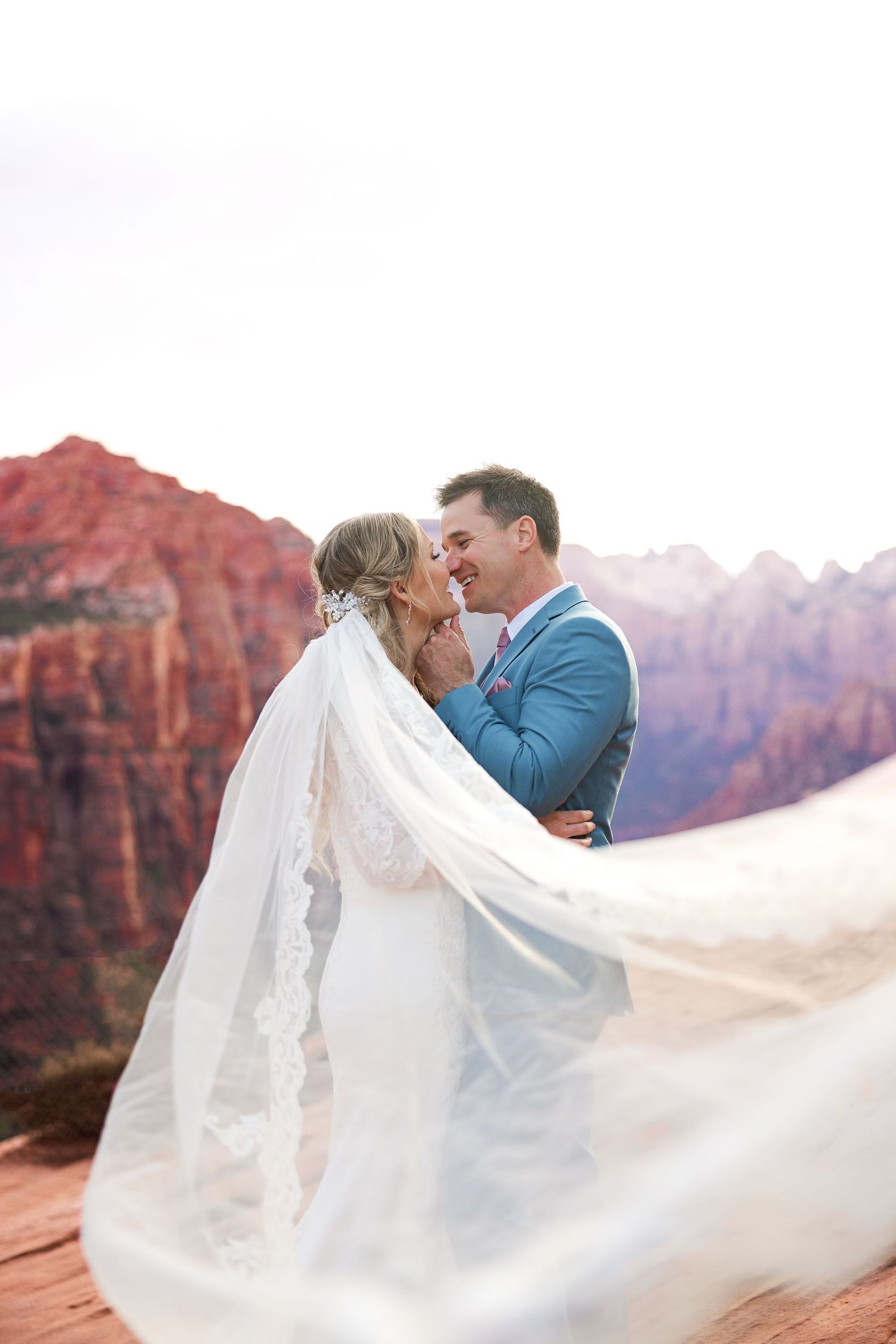A couple shares a romantic moment in a blue suit and white wedding dress against red rock cliffs at sunset.