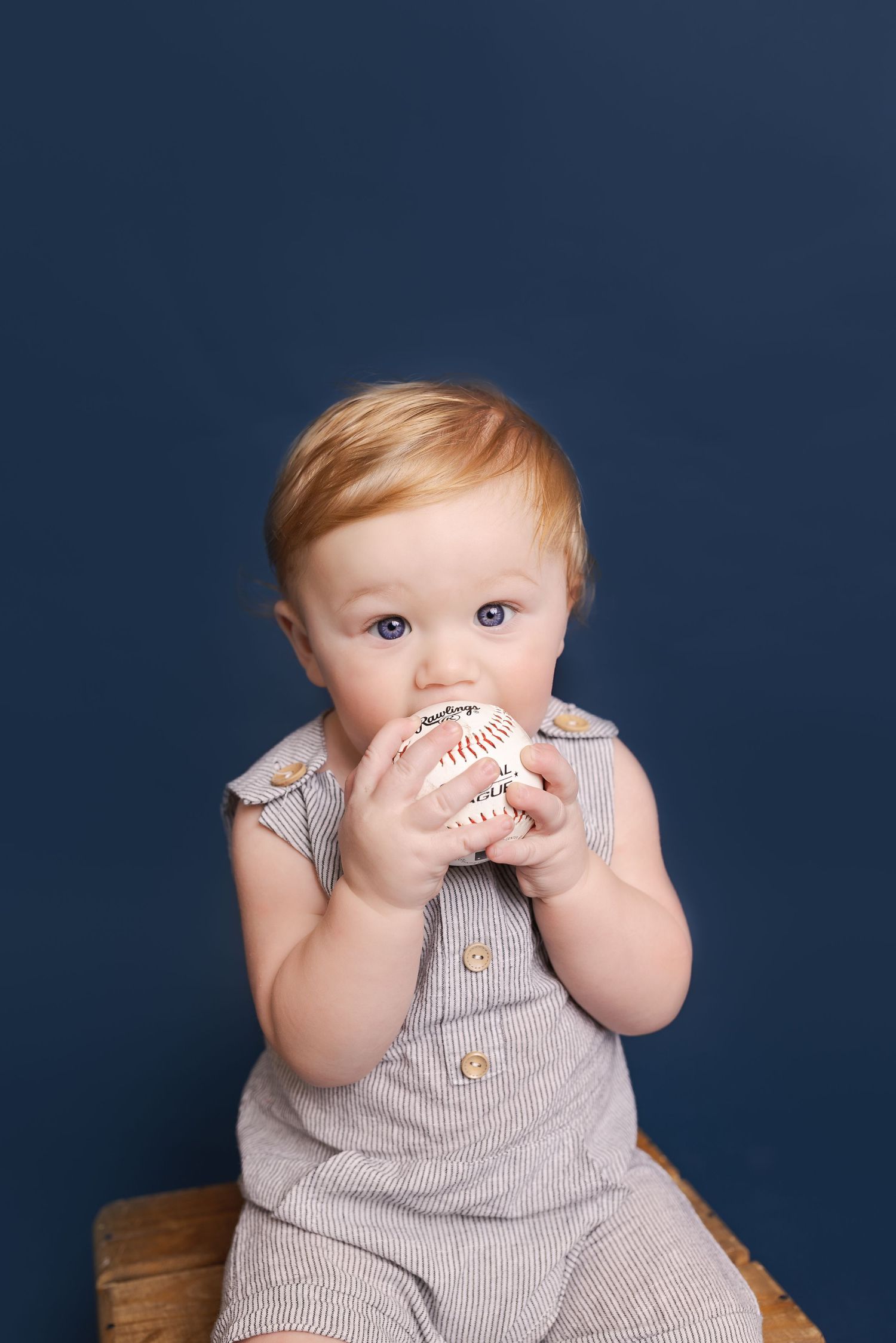Toddler in striped outfit holding baseball against dark blue backdrop.