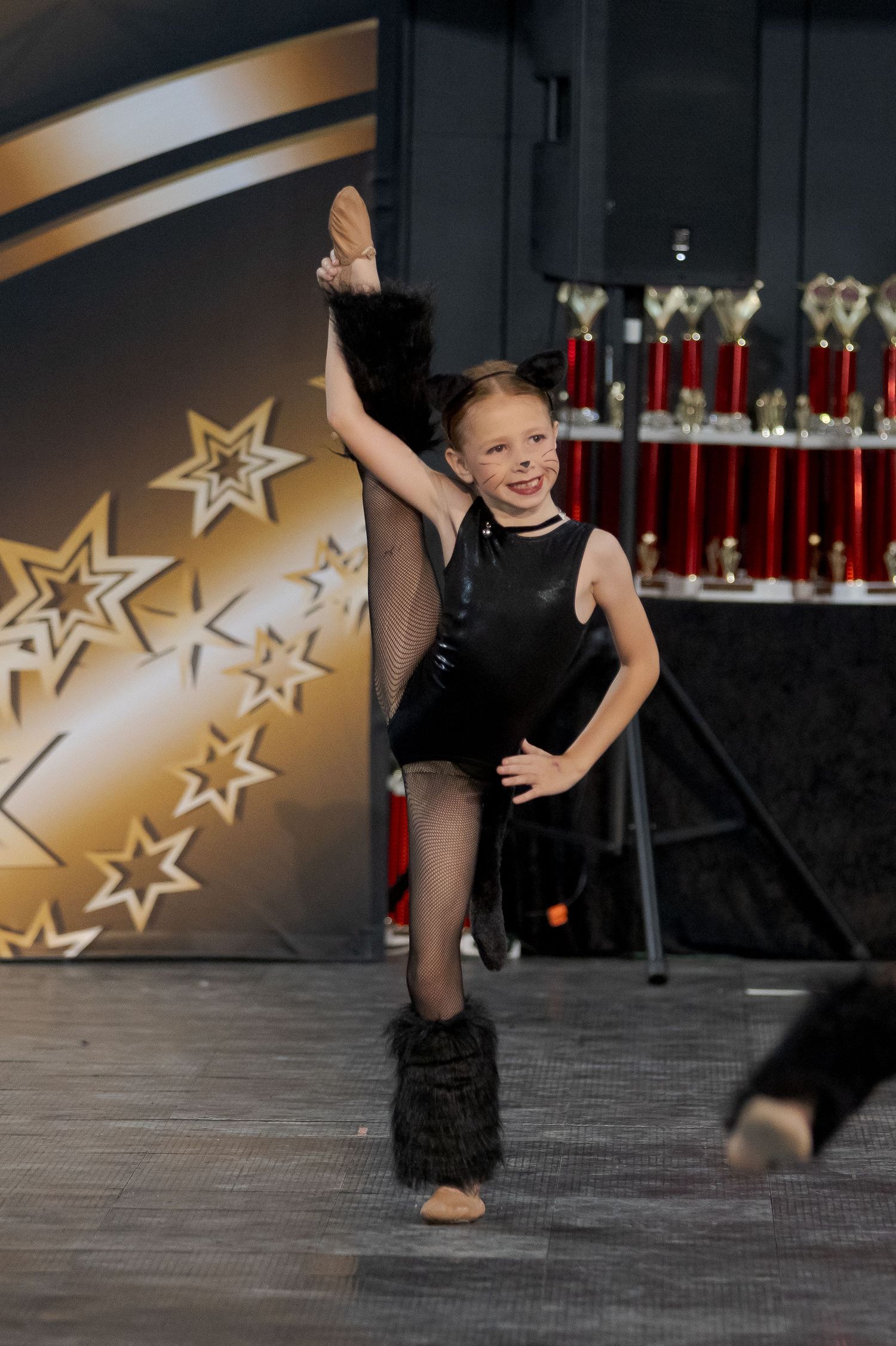 Young dancer in black leotard strikes a pose with leg extended on stage with star backdrop and trophies.