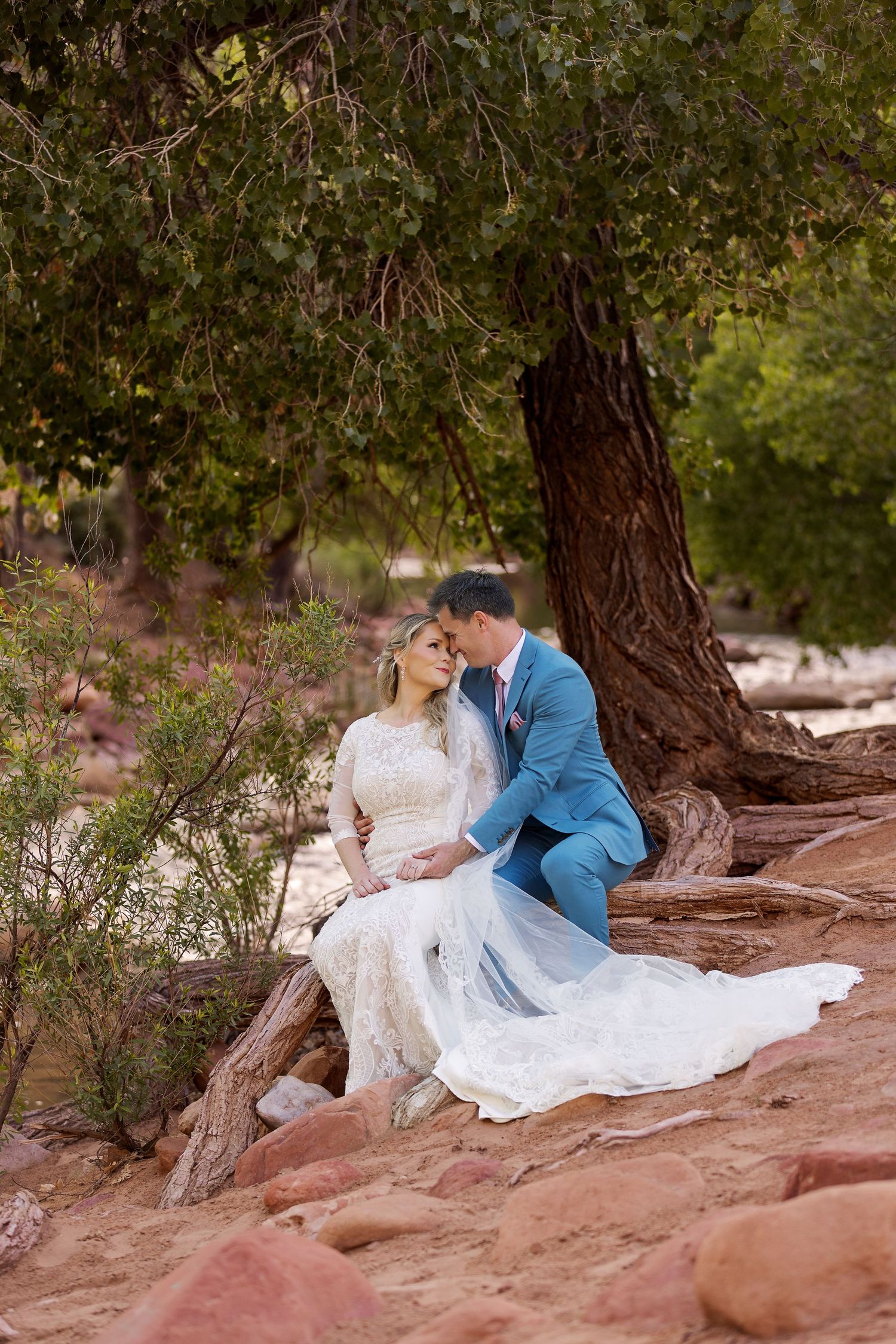Romantic couple in wedding attire sits together on red rocks surrounded by trees in a natural outdoor setting.