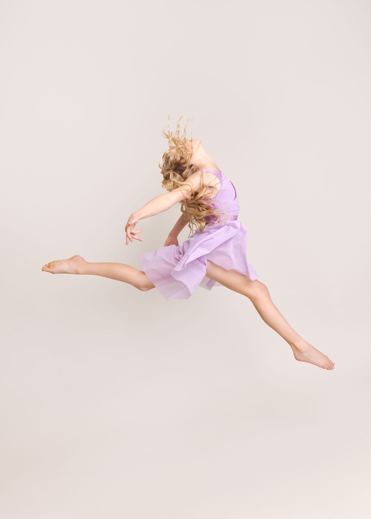 Dancer in lavender leotard performs graceful aerial leap against white backdrop.