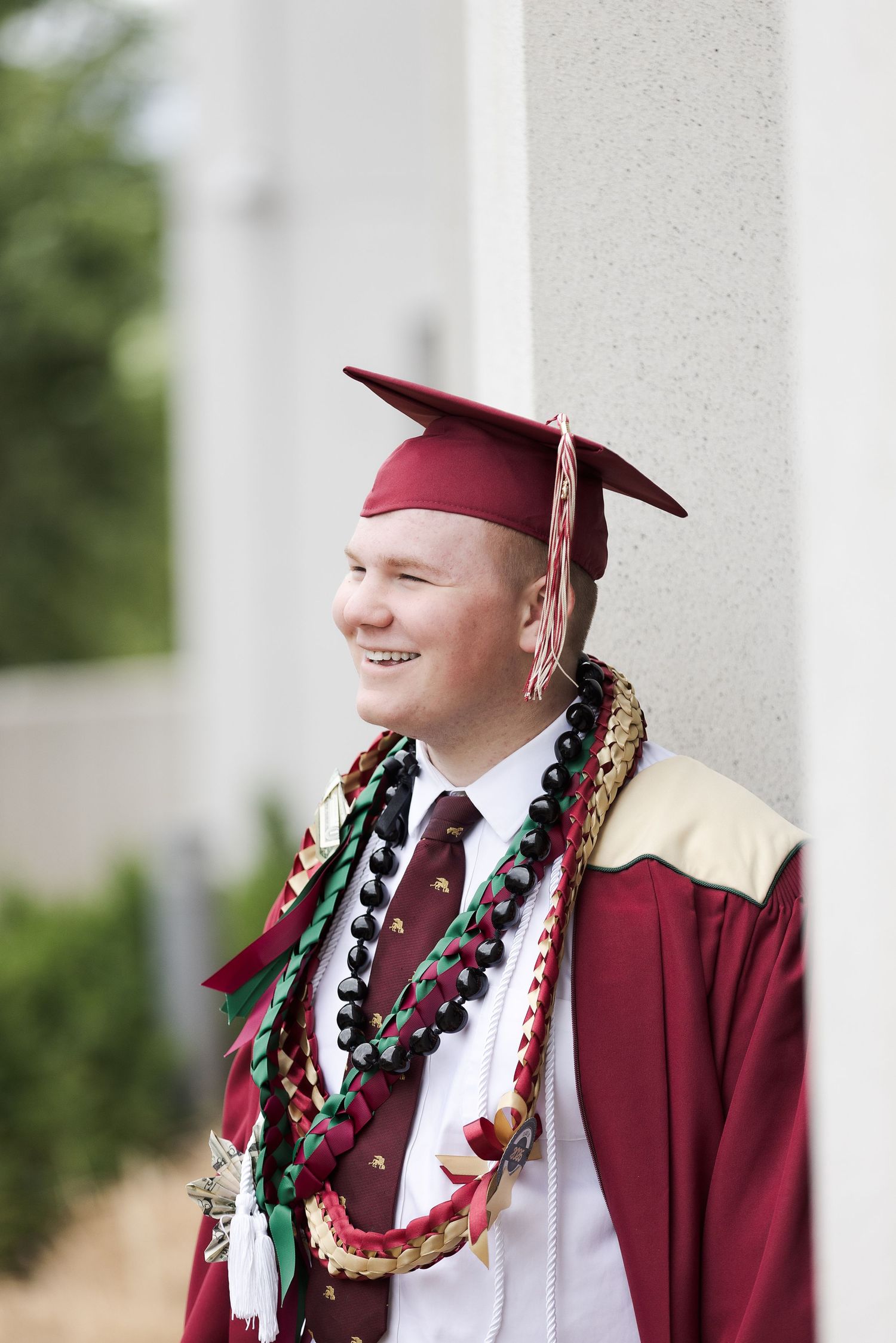 Smiling young man in maroon graduation cap and gown wearing leis, leaning against a white column outdoors.