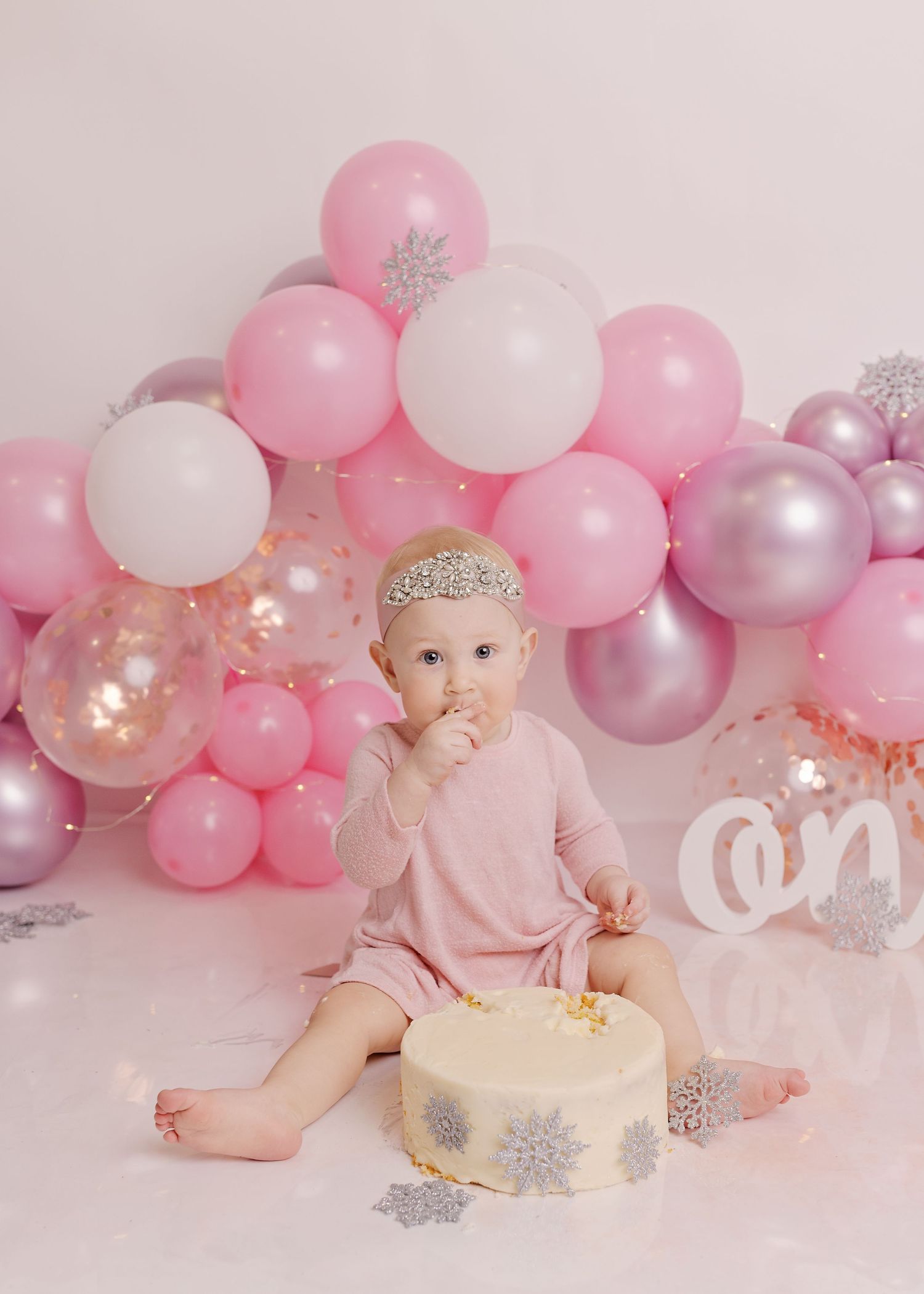 A baby in pink celebrates their cake smash photo session with pink and white balloons and sparkly snowflake decorations.