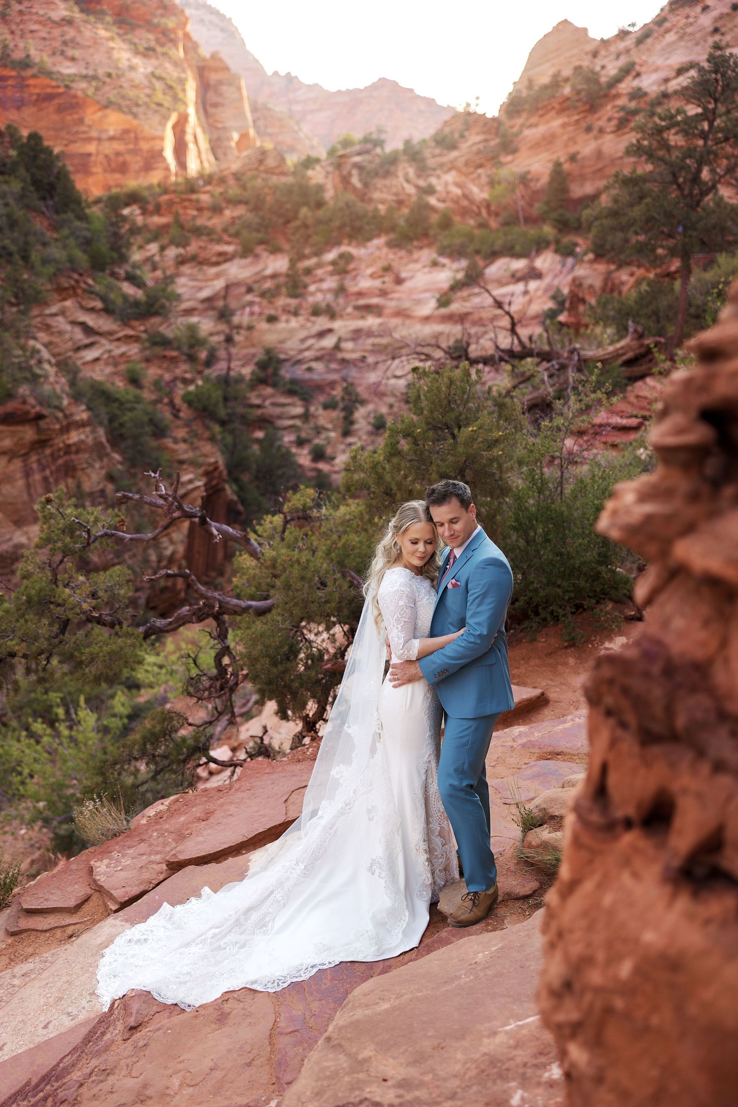 A couple shares an intimate moment on red sandstone cliffs among desert vegetation during sunset.