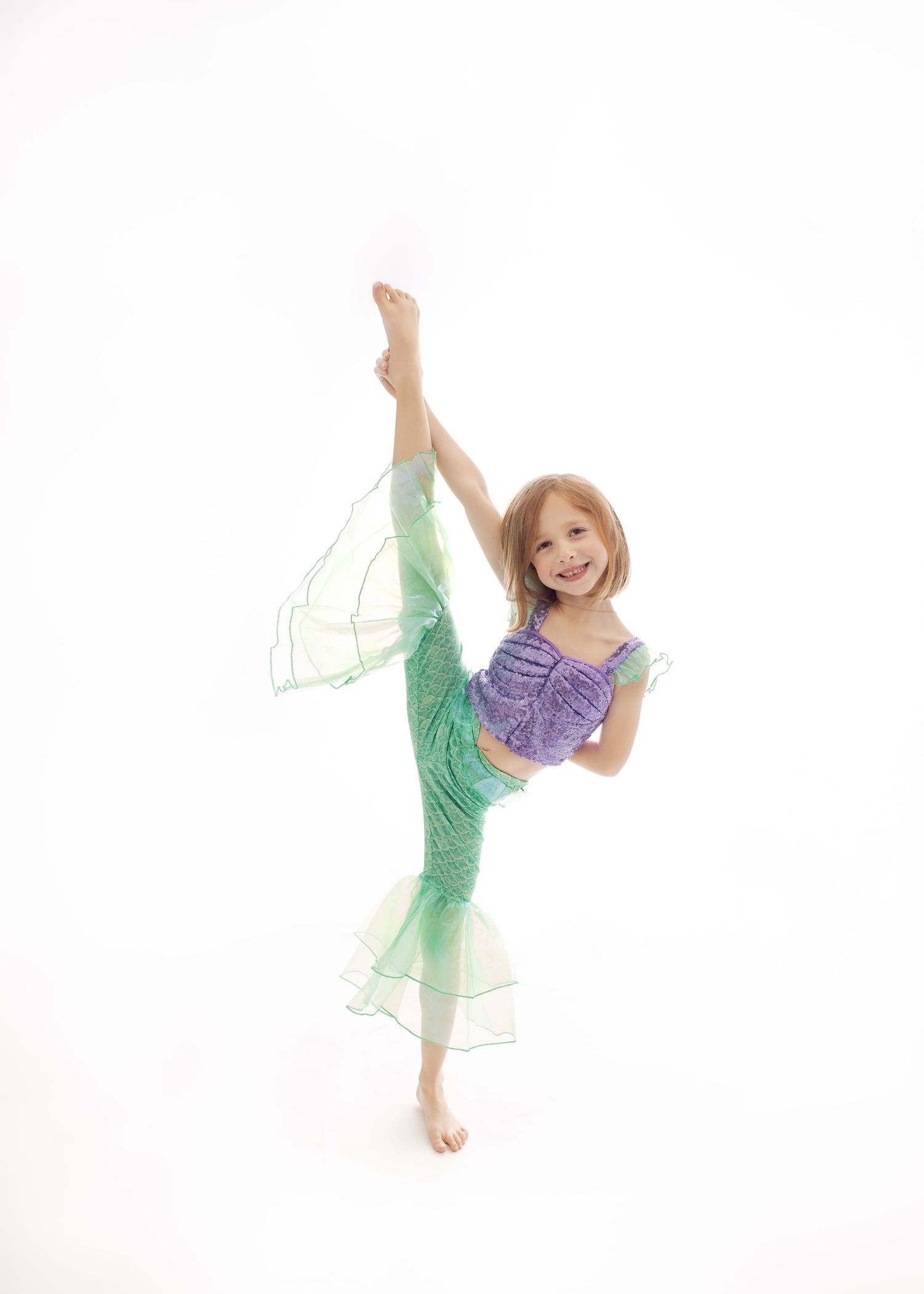 Young dancer in purple top and seafoam green costume performs graceful high kick against white background.