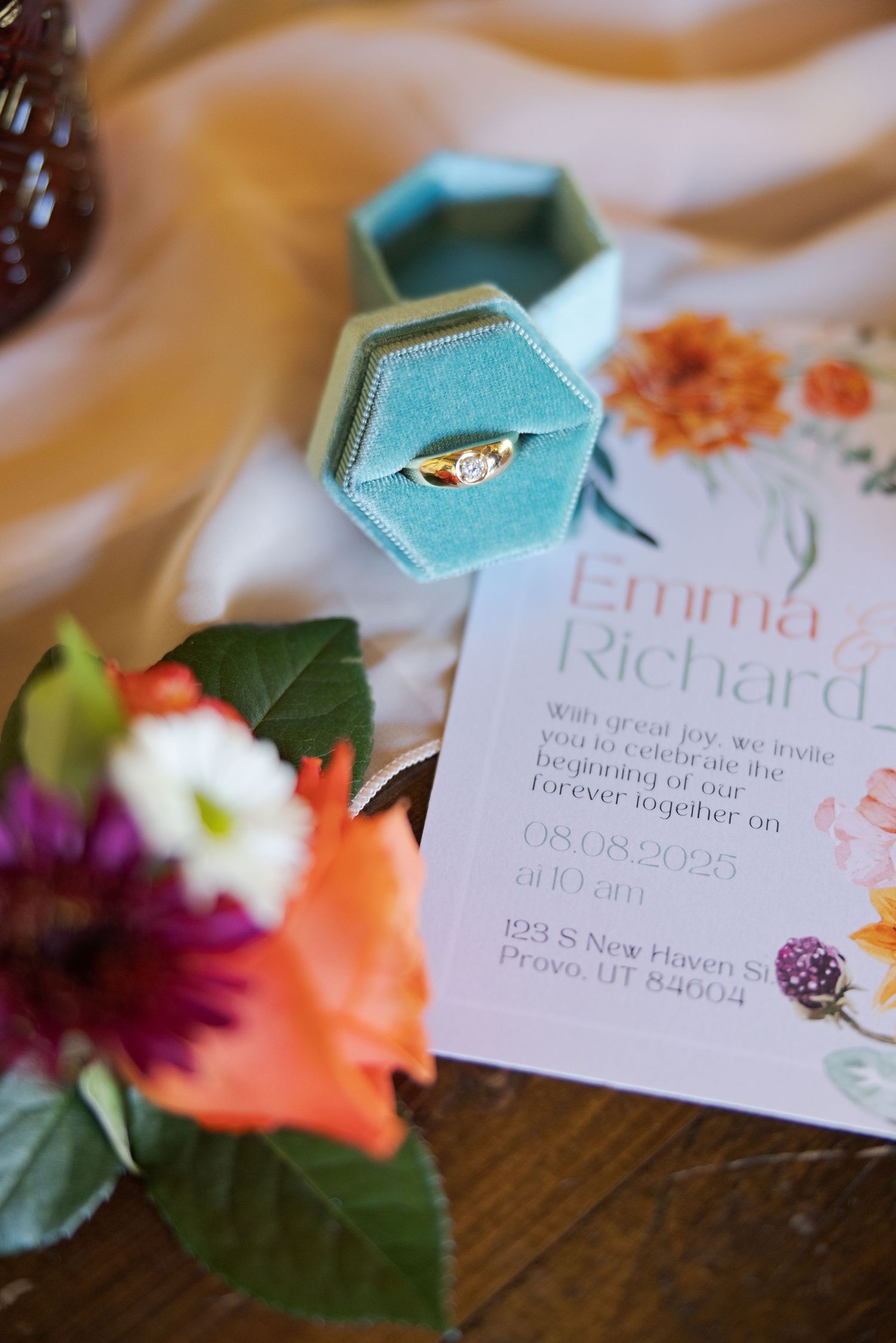 Wedding invitation details with turquoise ring box and orange floral accents on wooden surface.