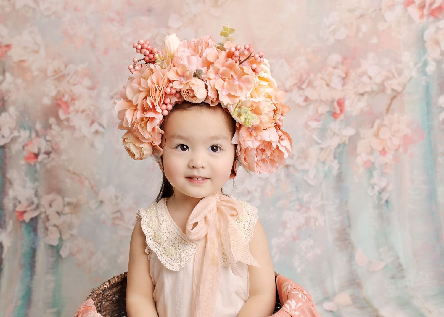 A dreamy portrait of a child wearing a peach-colored dress and floral crown against a pastel backdrop.
