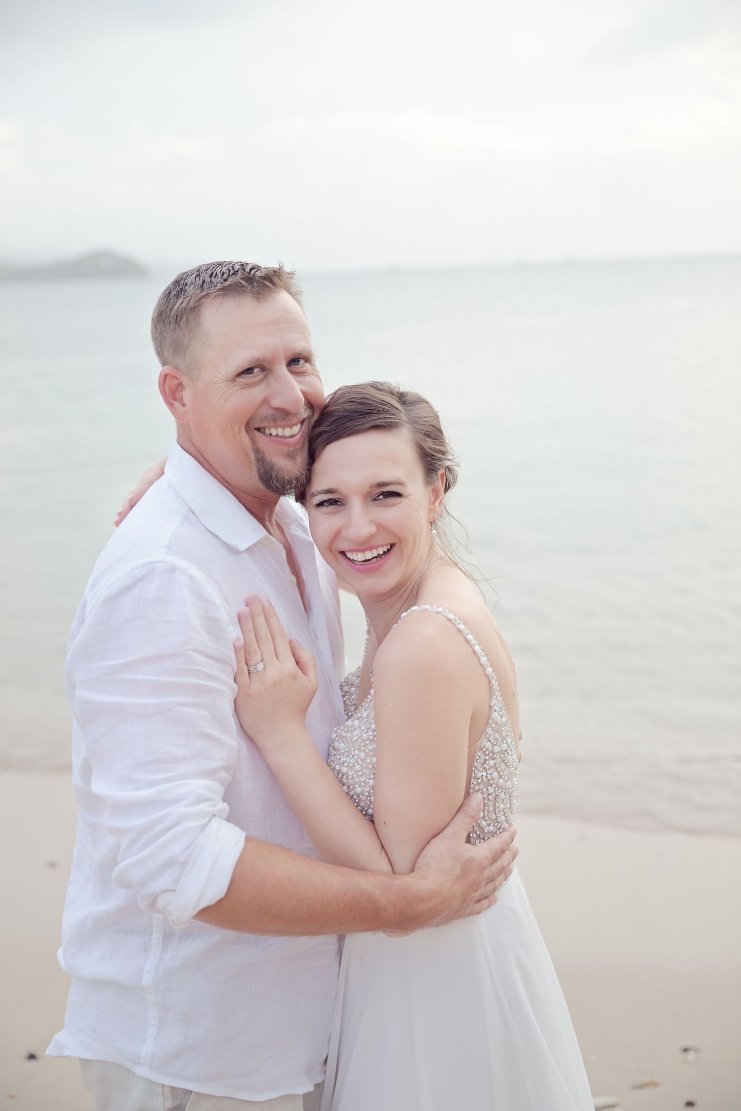 A happy couple embracing on a beach in white attire with soft lighting.