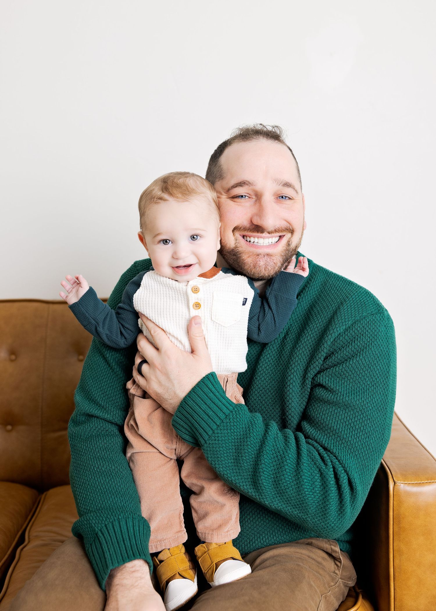 Smiling man in green sweater holds happy baby in white onesie and yellow shoes on brown leather couch.