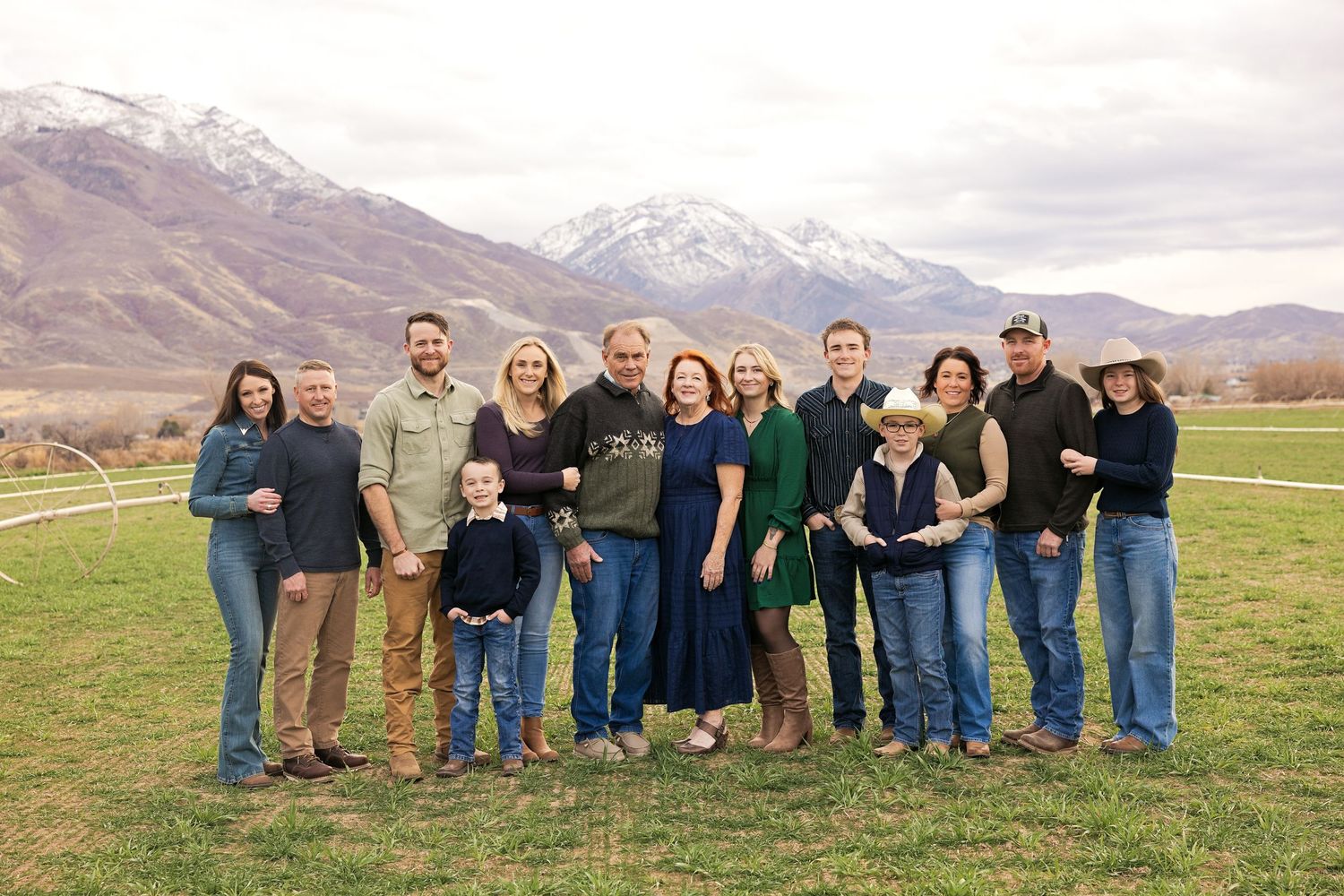 Large family group portrait outdoors with snow-capped mountains and green field backdrop.