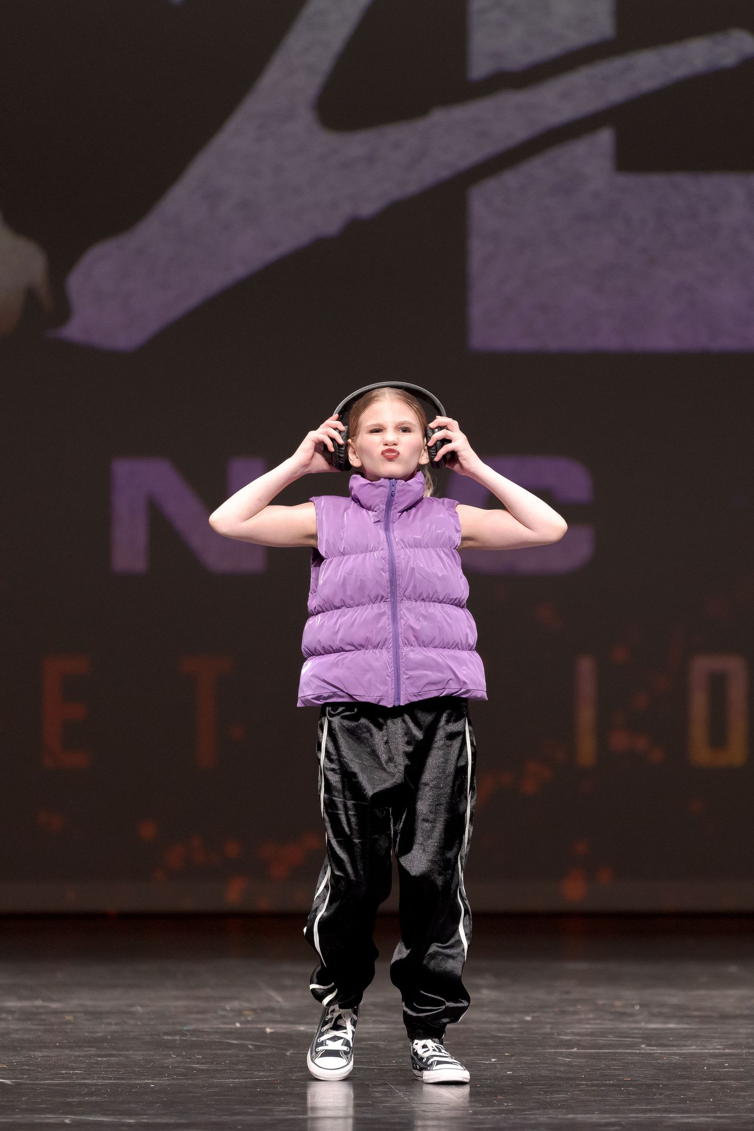Young performer in purple puffer vest and baggy pants wearing headphones during dance performance on stage.