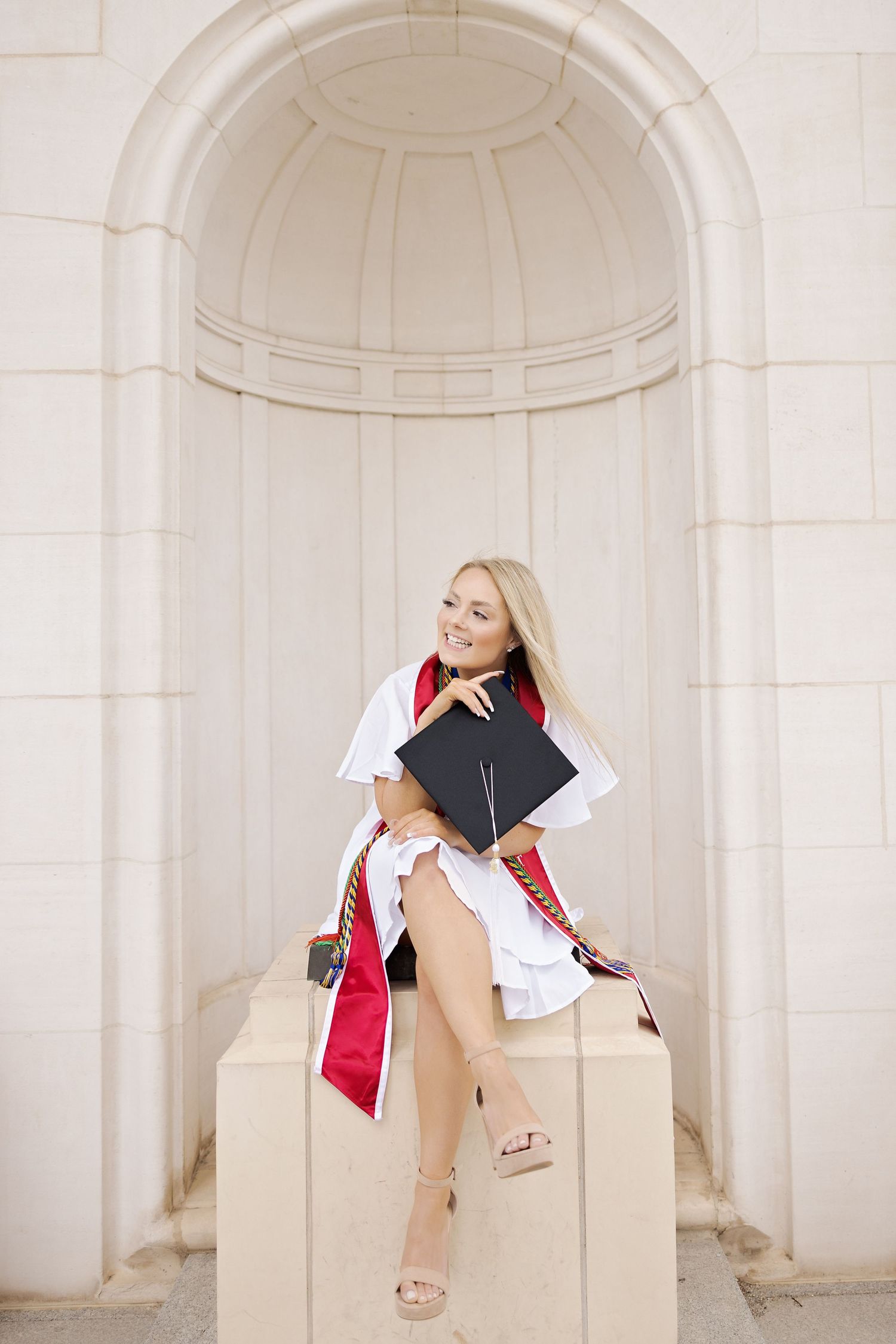Graduate in white dress and red stole posing with diploma in classic archway.