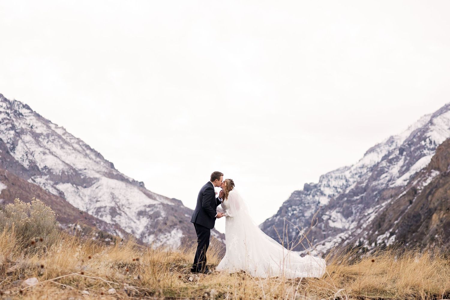 A romantic wedding photo shows a couple embracing on a hilltop with majestic snow-covered mountains behind them.