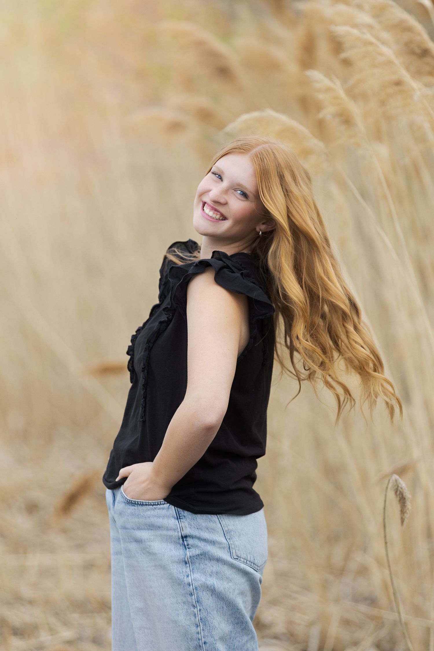 Smiling redhead girl in black ruffled top and jeans posing outdoors among tall golden grass.