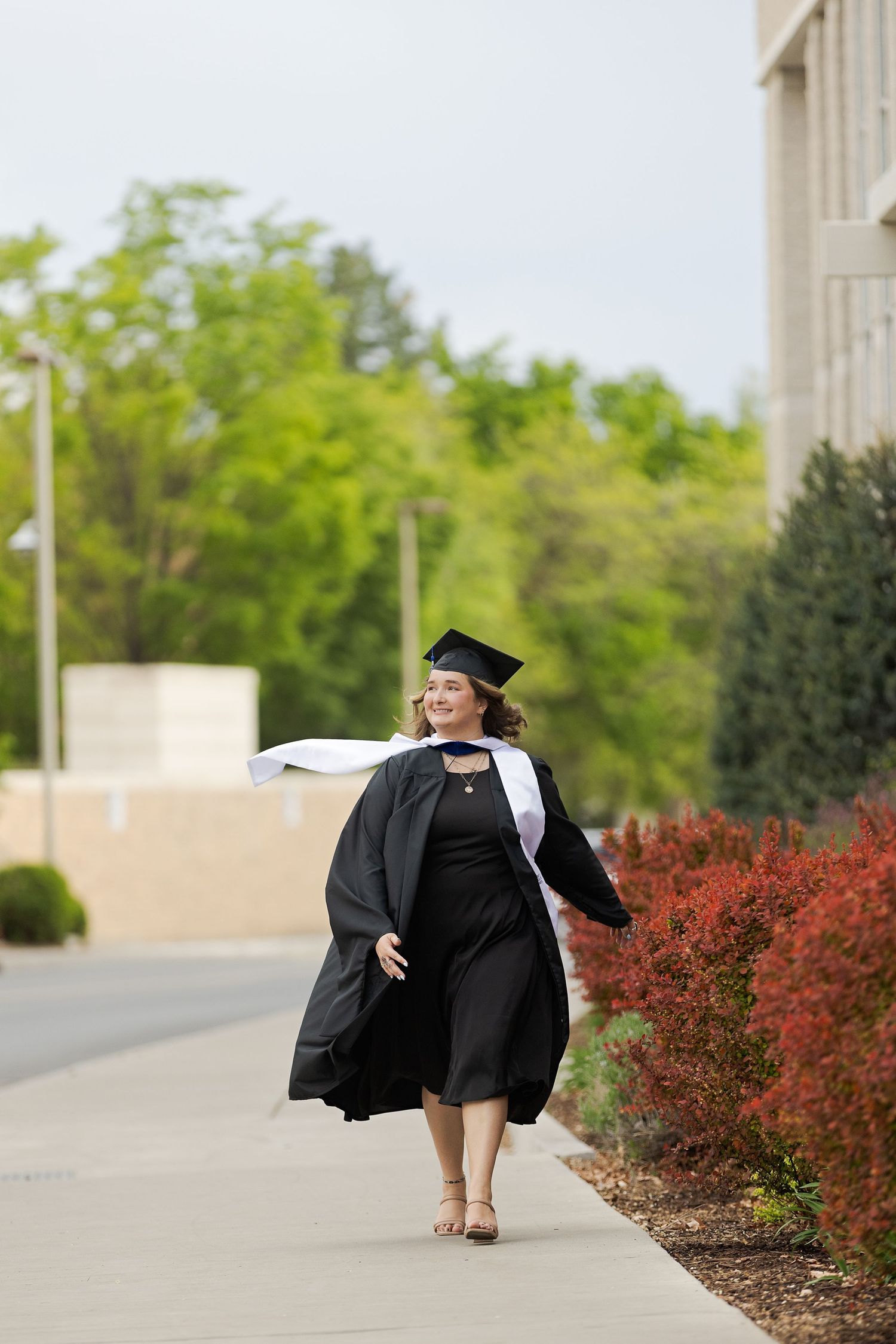 Smiling woman in black graduation gown and cap walks along campus path surrounded by lush greenery and red shrubs.