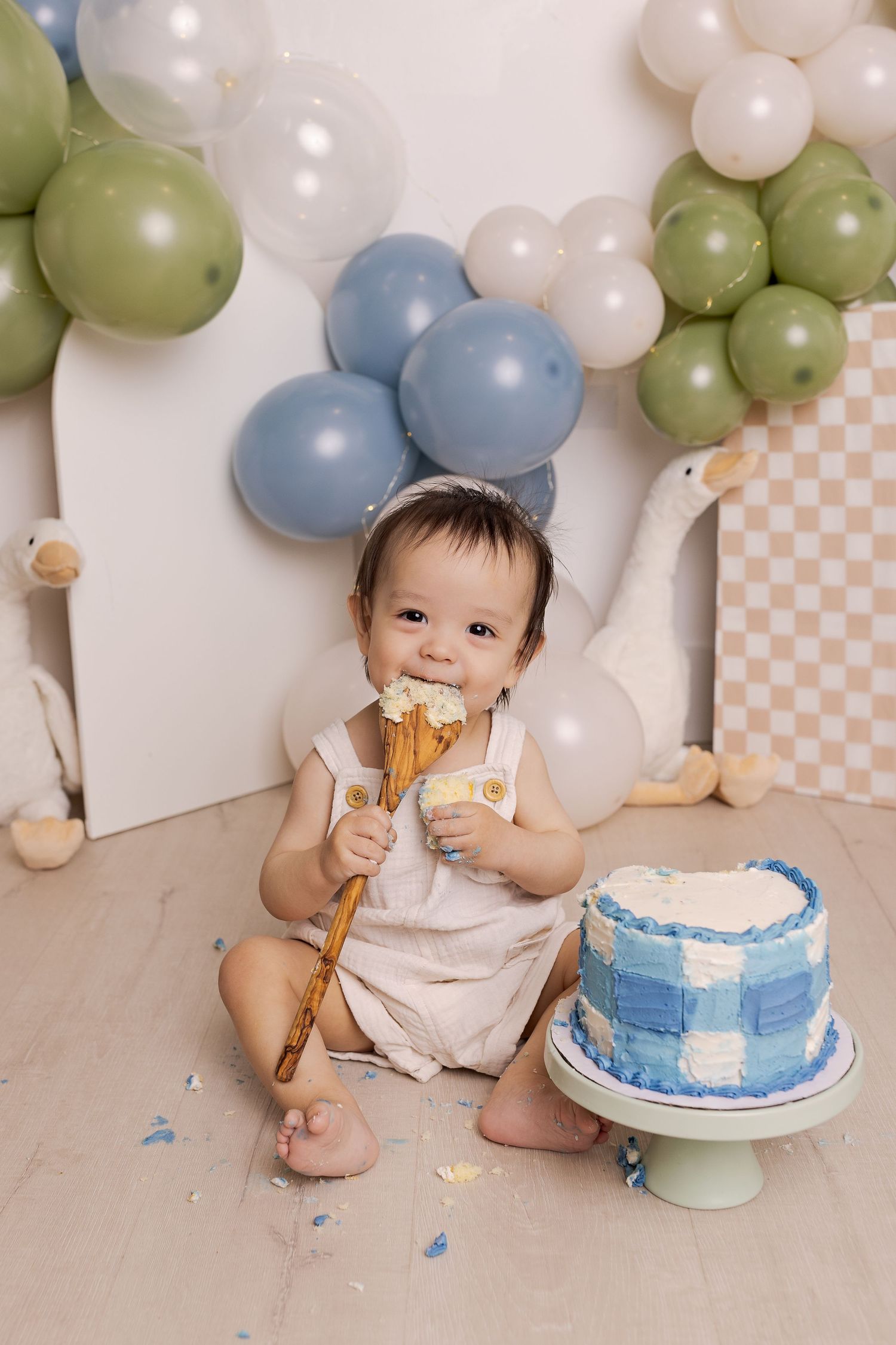 Baby in white romper smashing first birthday cake surrounded by blue, green, and white balloons at a cake smash photoshoot.