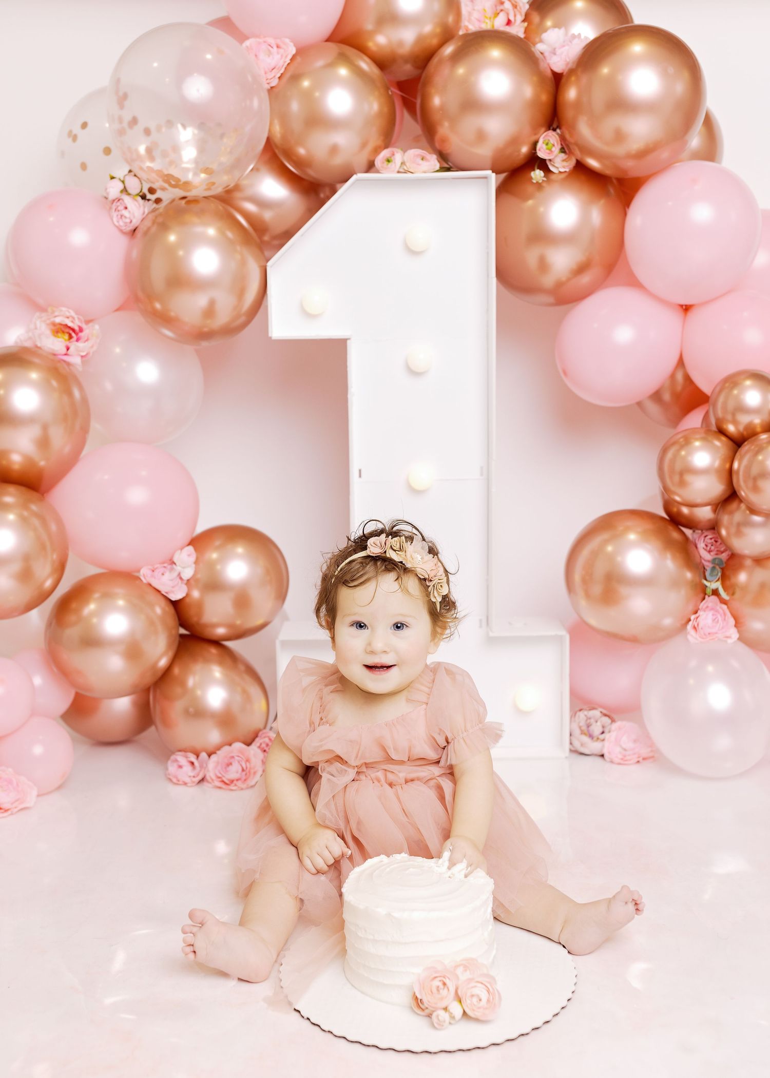 Pink and rose gold balloon arch with a large white number 1 centerpiece frames a cake smash setup in soft blush tones.