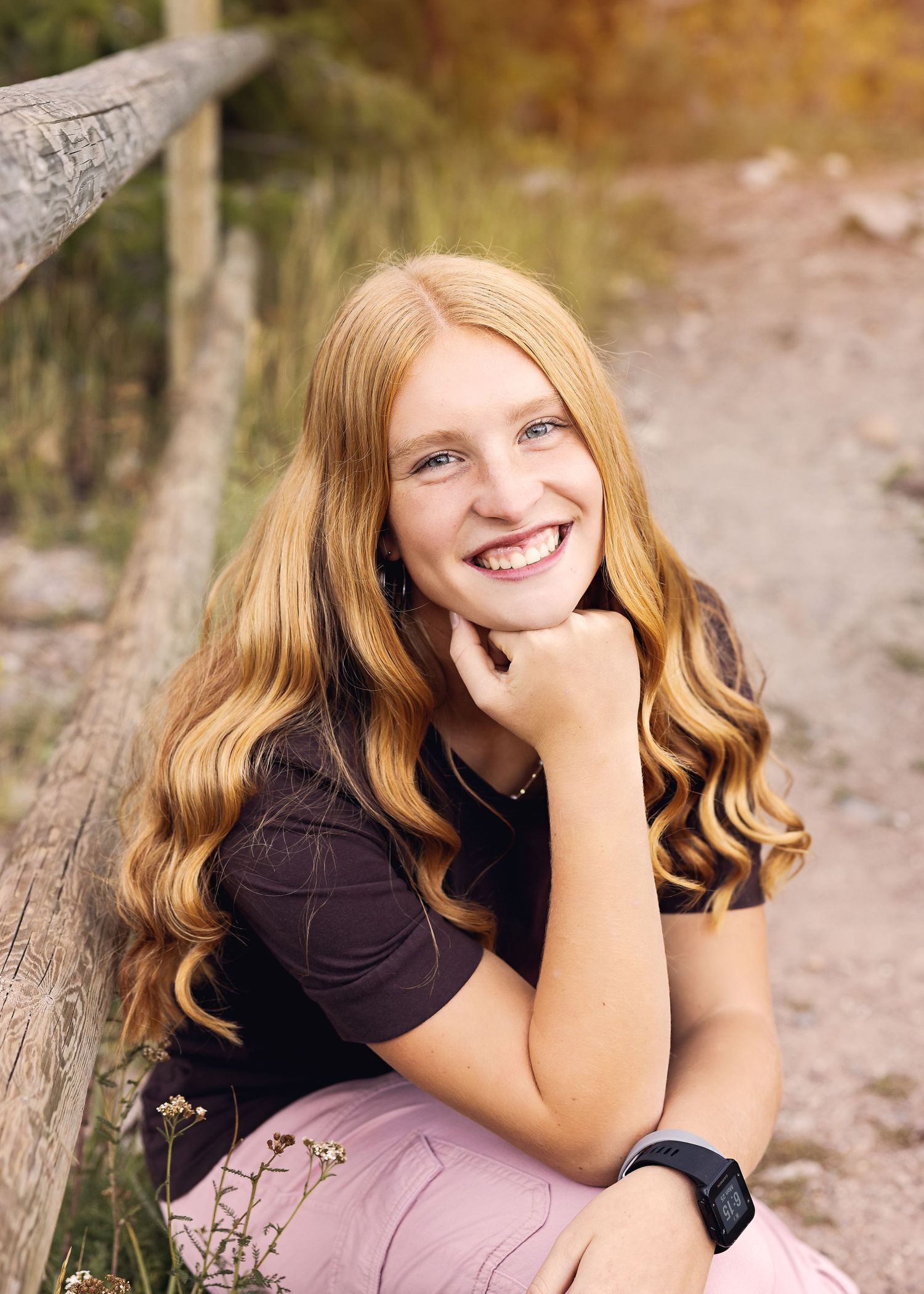 Young person with wavy blonde hair smiles while posing near wooden fence in autumn setting.